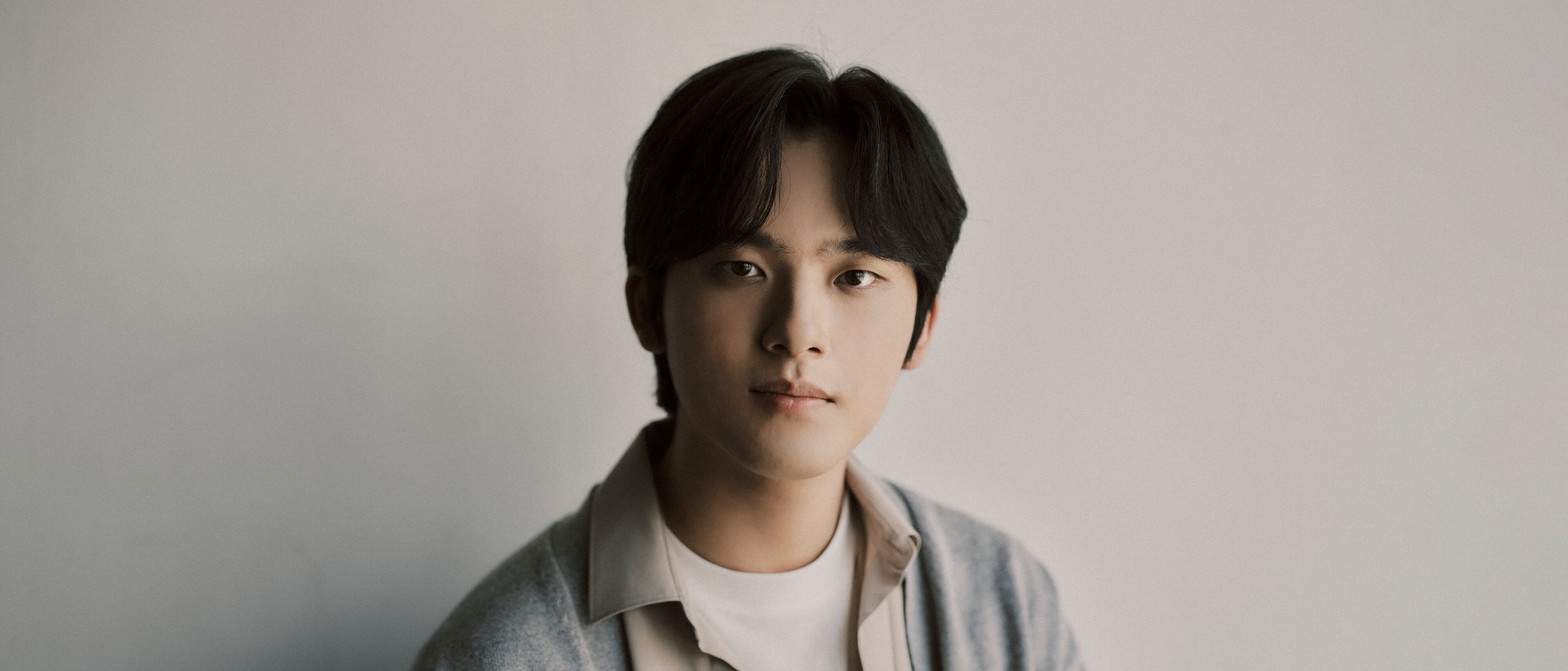 Yunchan Lim mit mittellangen dunklen Haaren, die ein weißes Hemd und eine graue Strickjacke trägt, sitzt vor einem schlichten hellen Hintergrund und schaut ruhig in die Kamera.