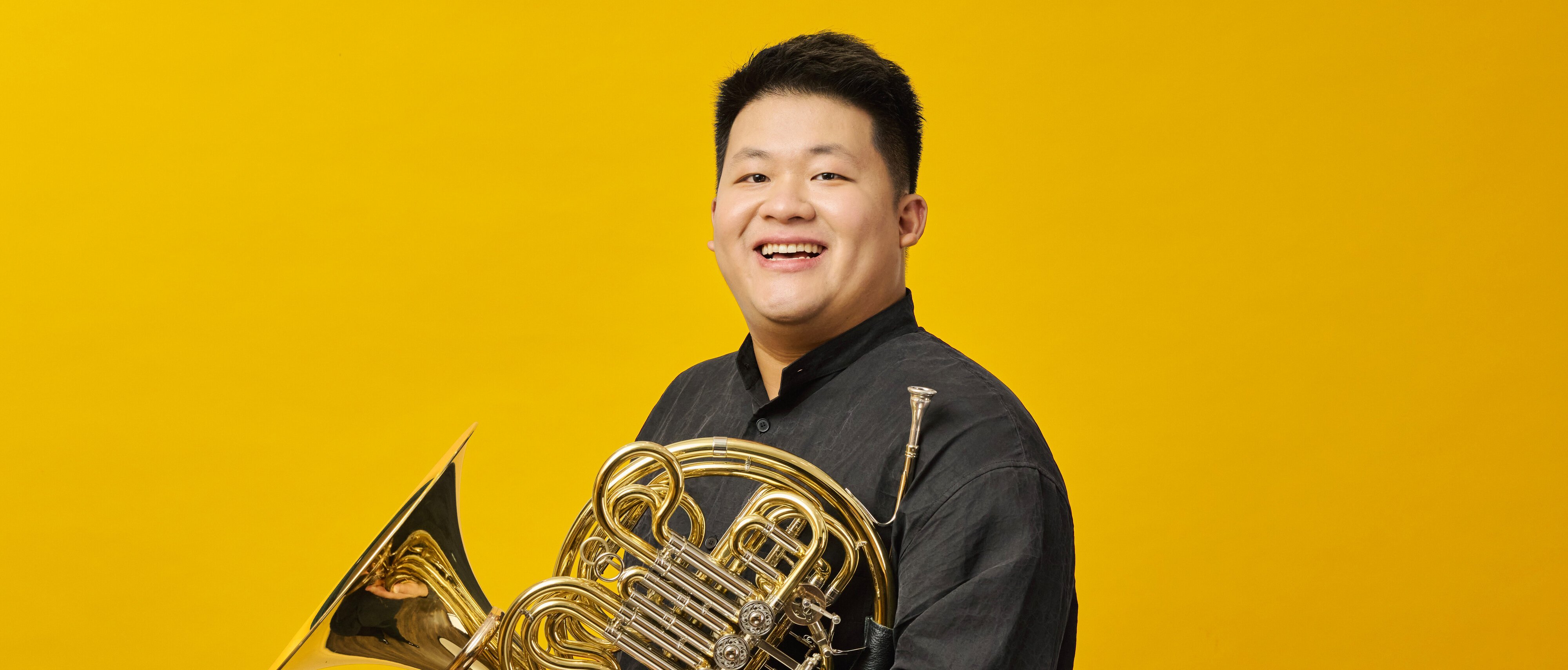 Yun Zeng in einem schwarzen Hemd hält ein Waldhorn vor einem leuchtend gelben Hintergrund.