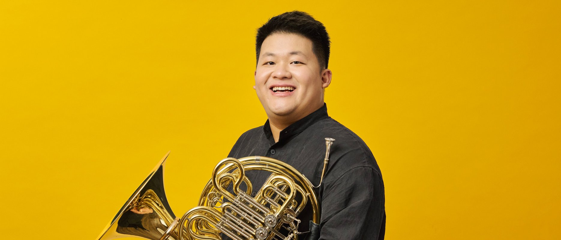 A smiling Yun Zeng wearing a dark shirt holds a French horn against a bright yellow background.