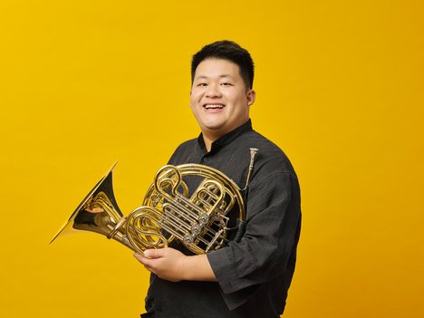 A smiling Yun Zeng wearing a dark shirt holds a French horn against a bright yellow background.