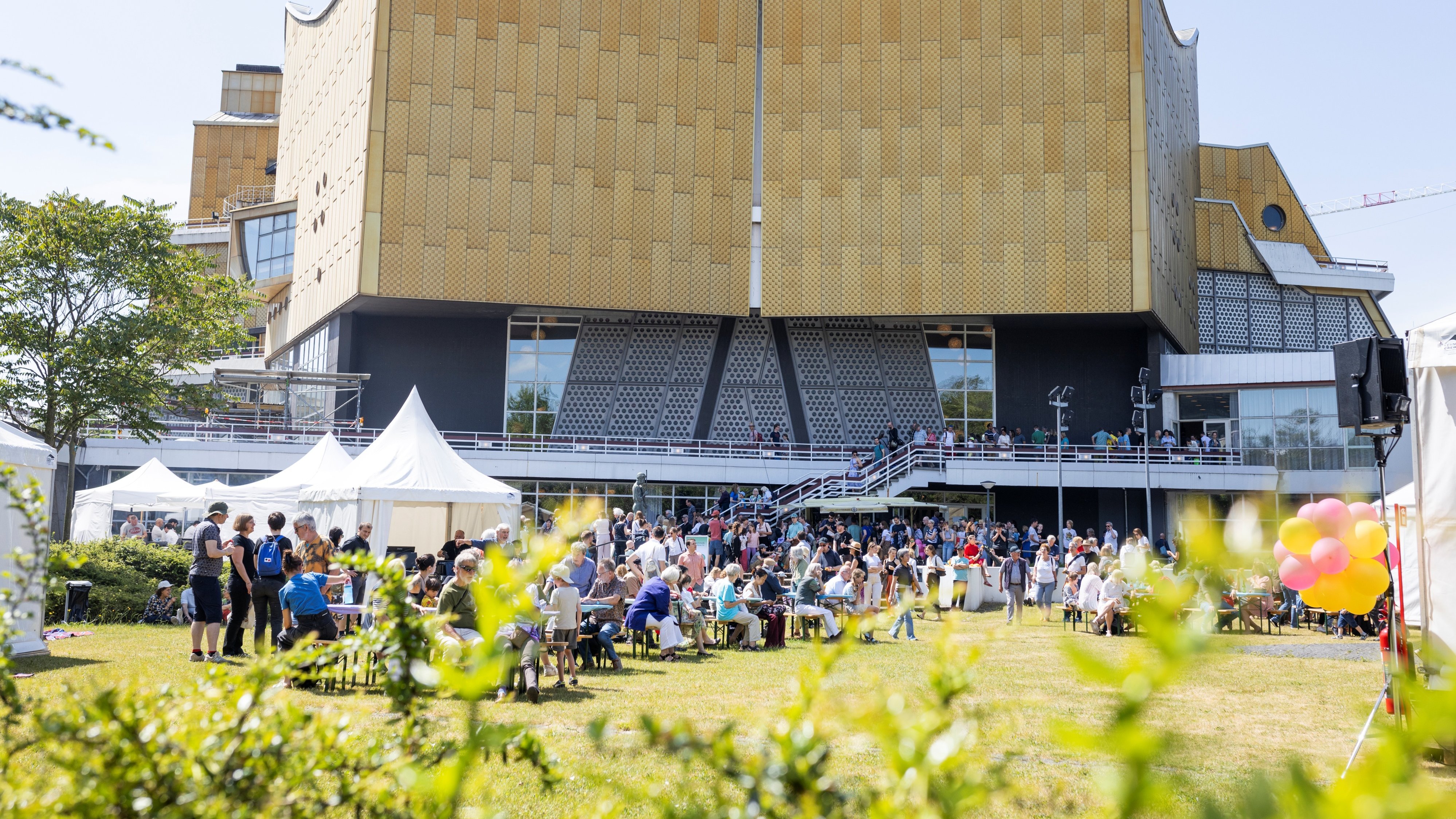 Vor der Philharmonie Berlin versammelt sich eine Menschenmenge, die an einem sonnigen Tag in der Nähe weißer Zelte sitzt und steht; rechts sind bunte Luftballons zu sehen.
