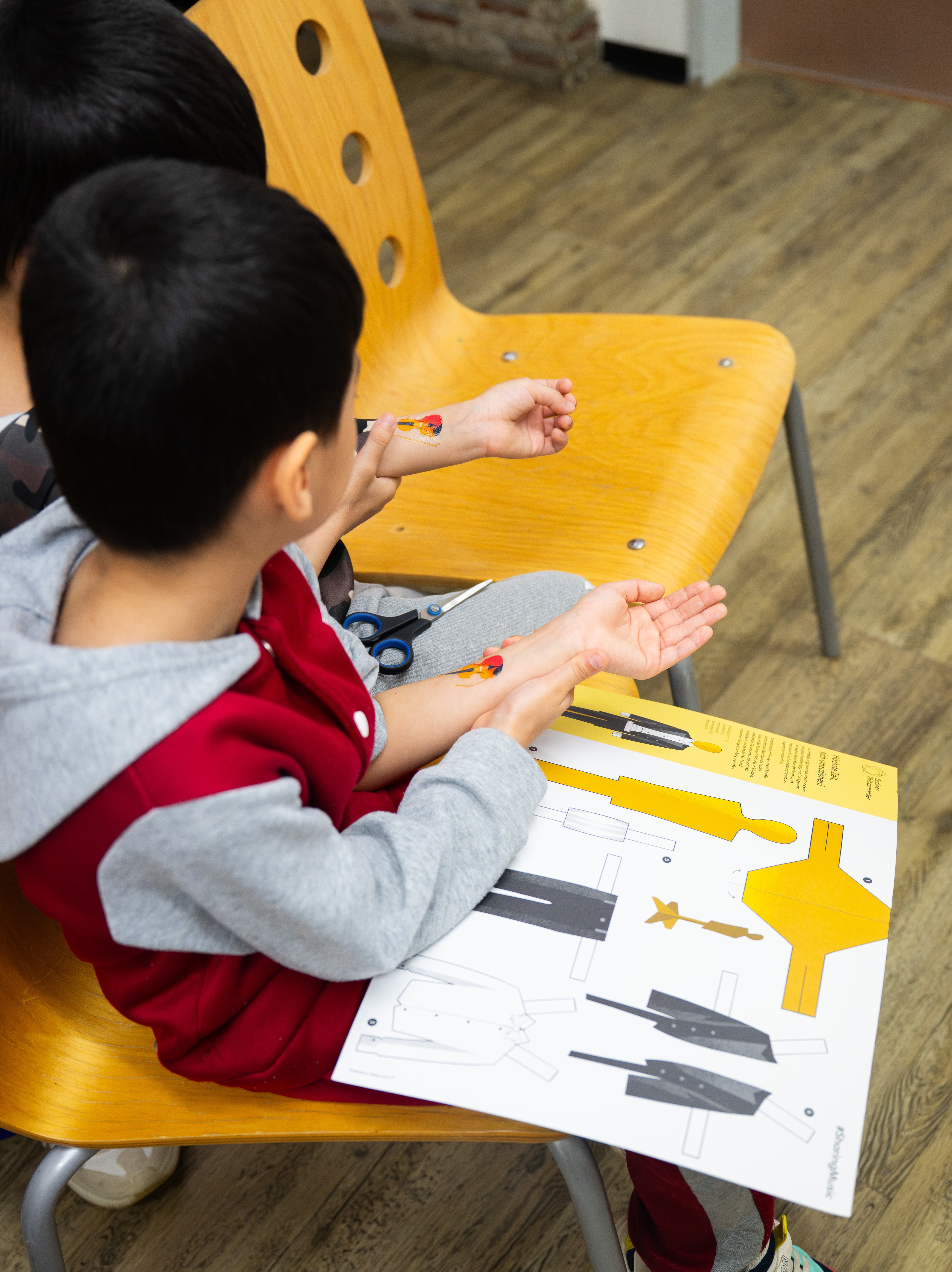 Two children are sitting on wooden chairs; one is holding a craft sheet and a pair of scissors. Both are looking at their wrists, where small violin motifs are stuck.