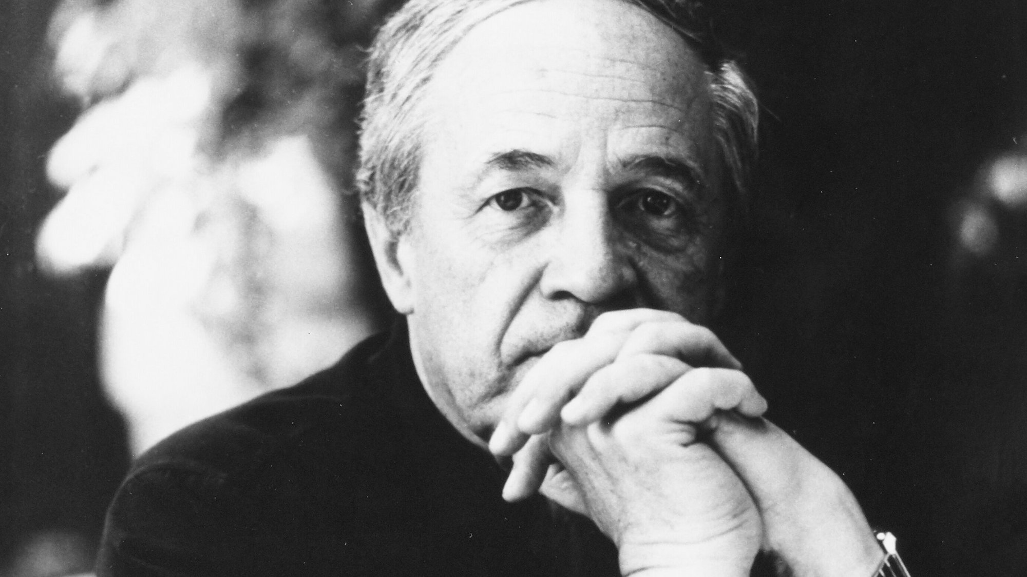 A black-and-white photo of Pierre Boulez with graying hair, wearing a dark short-sleeved shirt and a watch, sitting with his hands crossed in front of his face, gazing thoughtfully at the camera.