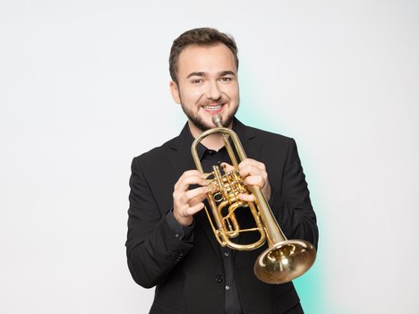 Eugenio Valle in a black suit holds a brass trumpet, standing against a plain white background.