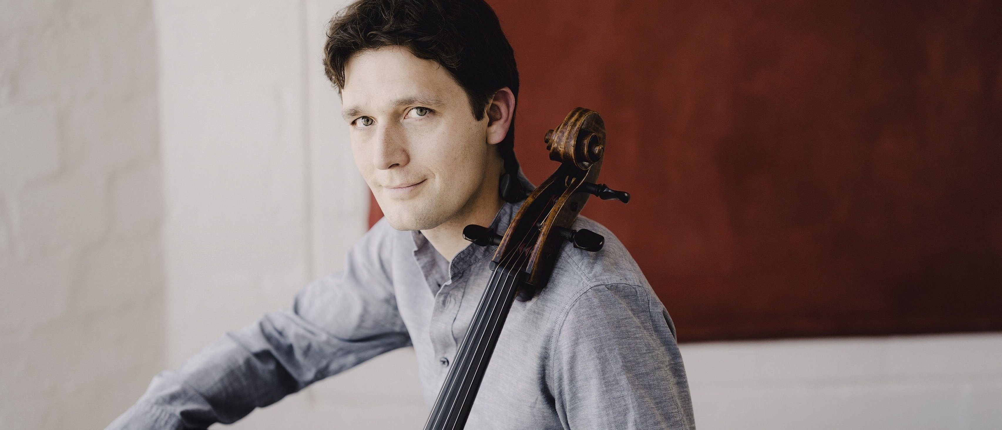 Junger Mann mit dunklem Haar in grauem Hemd, hält ein Cello und blickt freundlich in die Kamera. Er sitzt entspannt vor einer weißen Wand mit rotem Hintergrundelement.