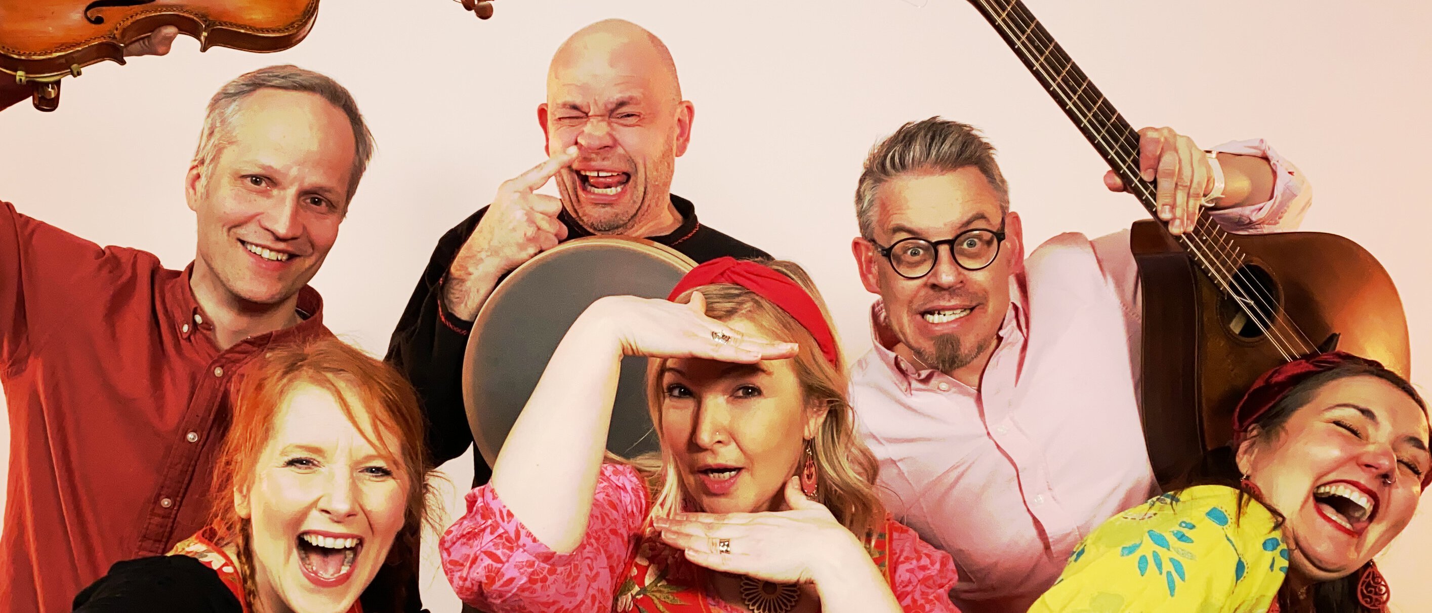 Six joyful adults in colorful clothing pose playfully with musical instruments, including a violin and guitar, against a light background. They are smiling and gesturing energetically toward the camera.
