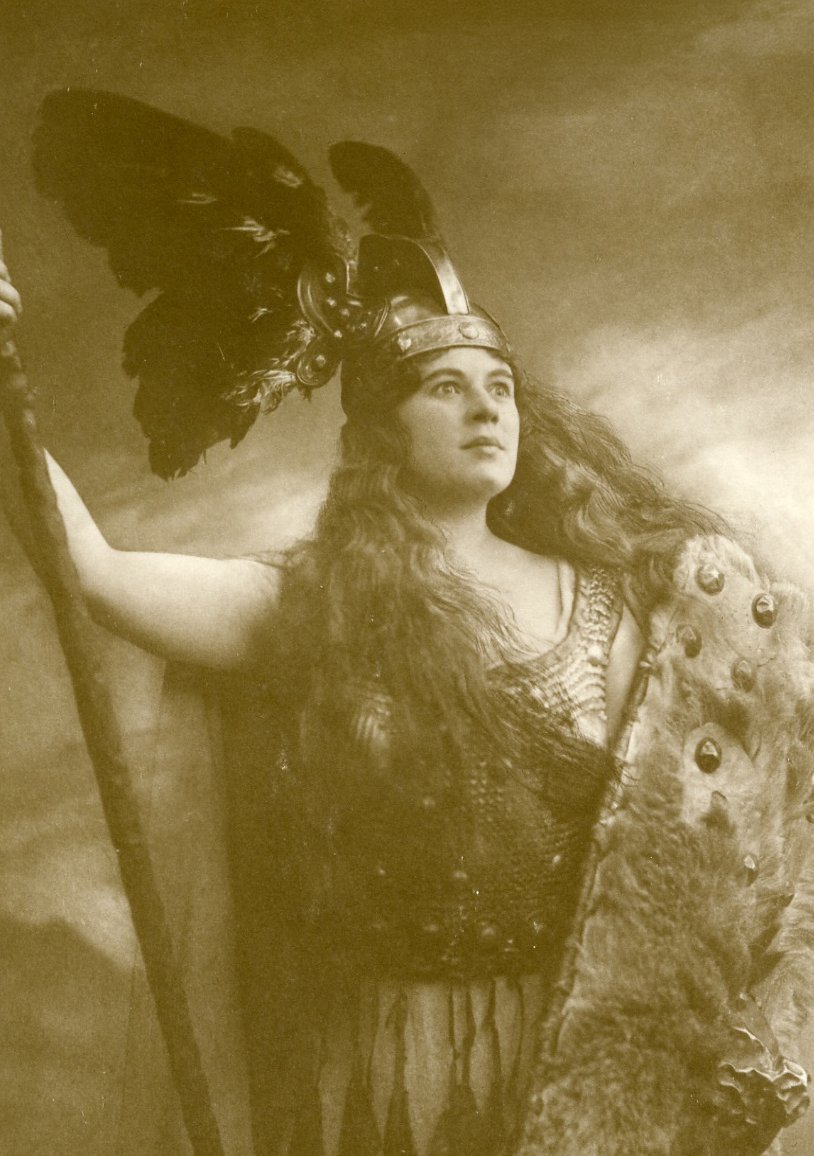A woman in a winged helmet, chainmail, and fur cape holds a staff, gazing upward with a serious expression in this sepia-toned, dramatic portrait inspired by Norse mythology.