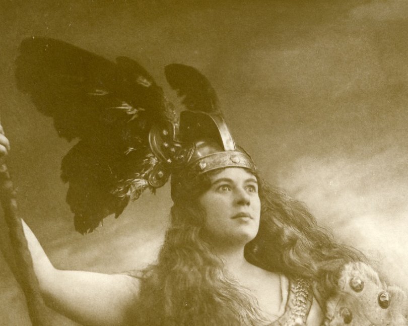 A woman in a winged helmet, chainmail, and fur cape holds a staff, gazing upward with a serious expression in this sepia-toned, dramatic portrait inspired by Norse mythology.