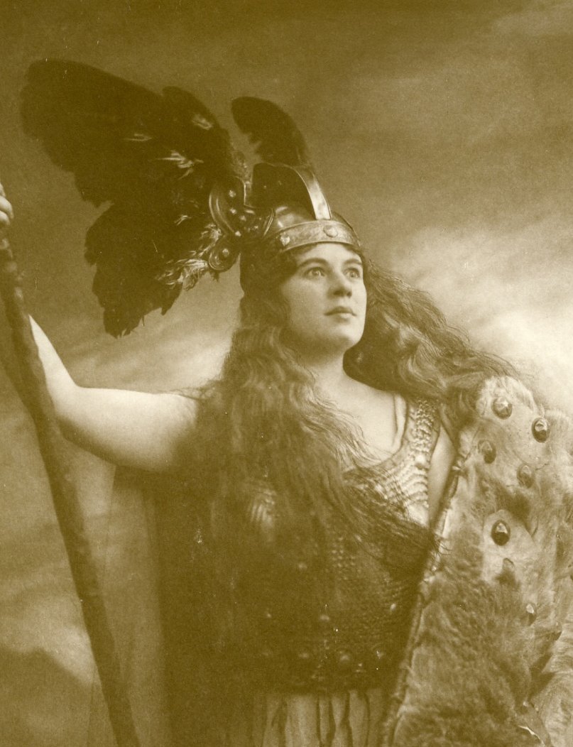 Eine Person in einem Kostüm mit einem geflügelten Helm, einer metallischen Rüstung und einem großen Schild hebt einen Arm und scheint eine Figur aus der Mythologie oder einer Oper darzustellen. Das Bild ist in Schwarz-Weiß gehalten.