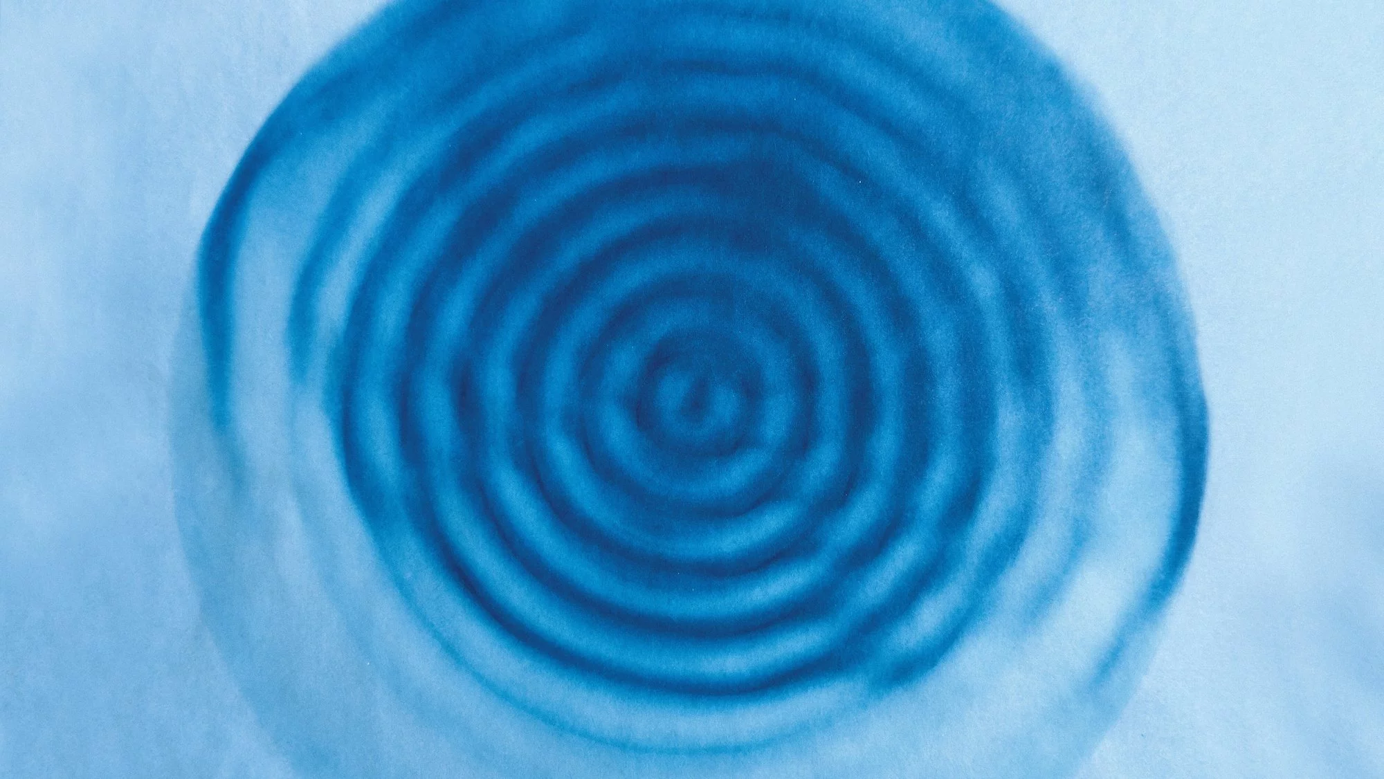Blue concentric circles radiate outward on a light blue background, resembling ripples in water or a topographic map. The image has a soft, abstract, and watercolor-like quality.