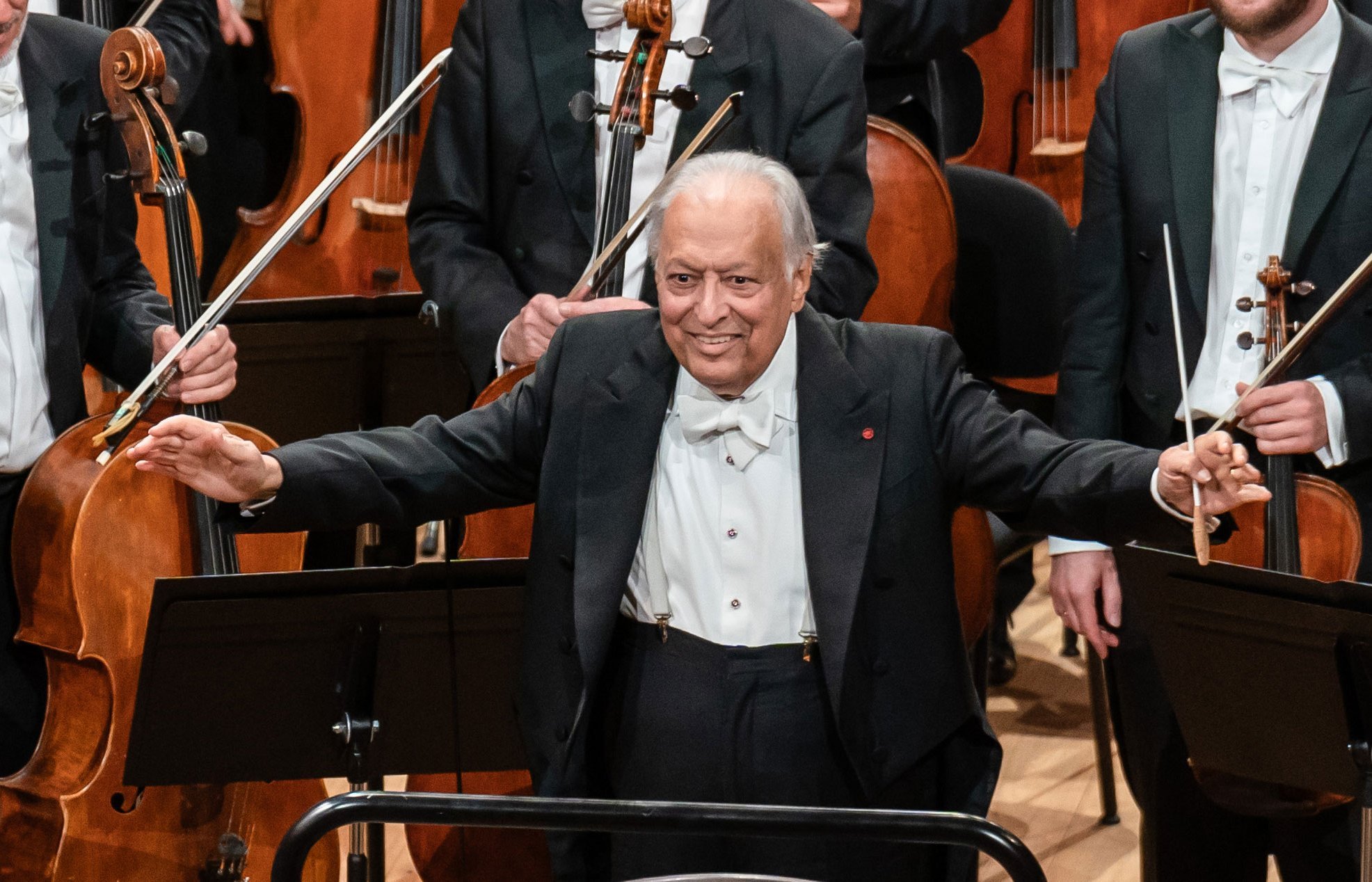 Ein älterer männlicher Dirigent in förmlicher Kleidung steht lächelnd mit erhobenen Armen, einen Taktstock haltend, vor einem Orchester mit Musikern und Streichinstrumenten hinter ihm.