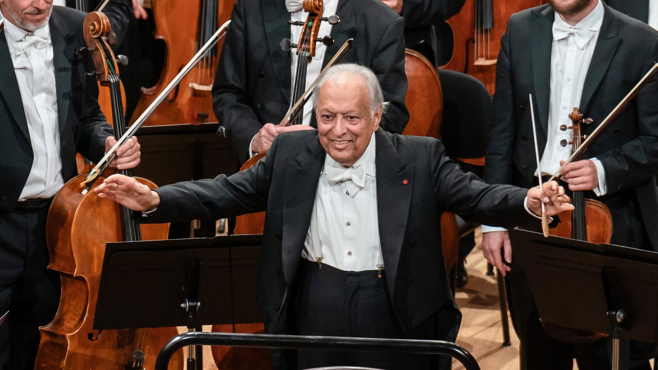 Ein älterer männlicher Dirigent in förmlicher Kleidung steht lächelnd mit erhobenen Armen, einen Taktstock haltend, vor einem Orchester mit Musikern und Streichinstrumenten hinter ihm.