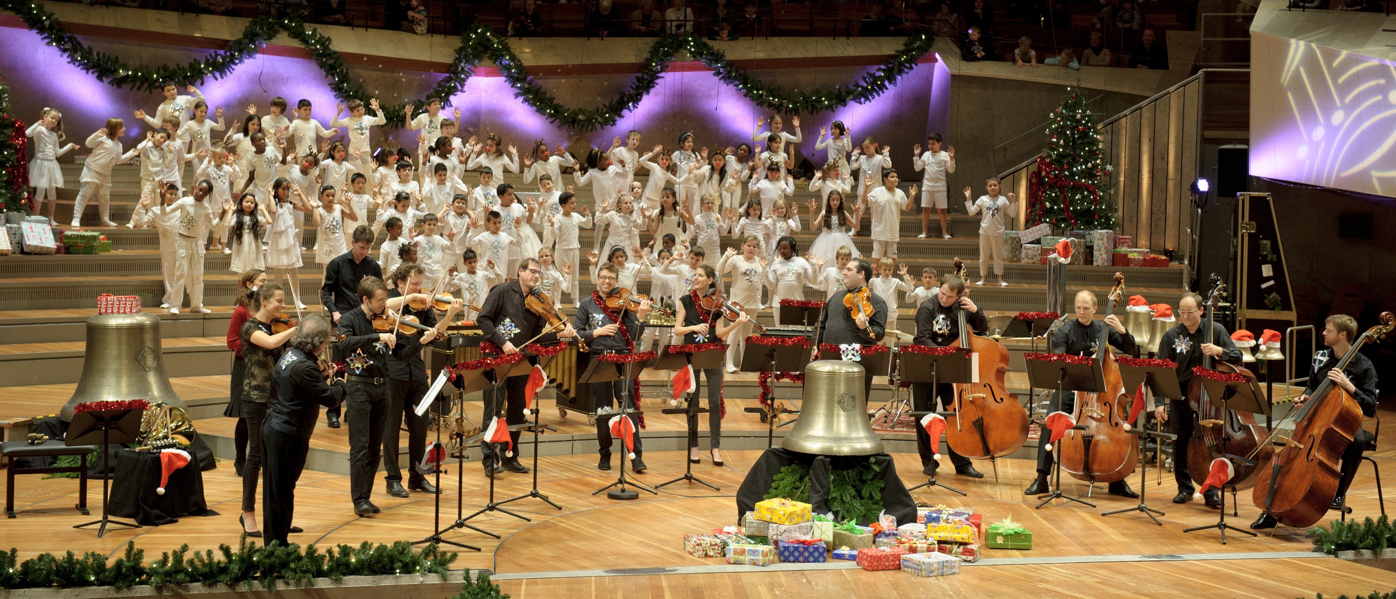 Ein großes Orchester und ein Chor treten auf einer mit Weihnachtsbäumen, Girlanden und Geschenken geschmückten Bühne auf, während Musiker Streichinstrumente und Glocken spielen. Der weiß gekleidete Chor steht hinter dem Orchester.