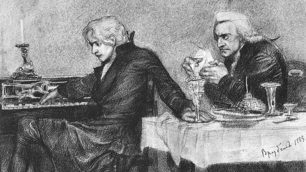 Mozart und Salieri in dunkler Kleidung sitzen in einem schwach beleuchteten Raum. Mozart spielt ernst Klavier, während Salieri an einem Tisch sitzt, intensiv Mozart anstarrt und ein Blatt Papier in der Hand hält. Daneben stehen Essens- und Weingläser.