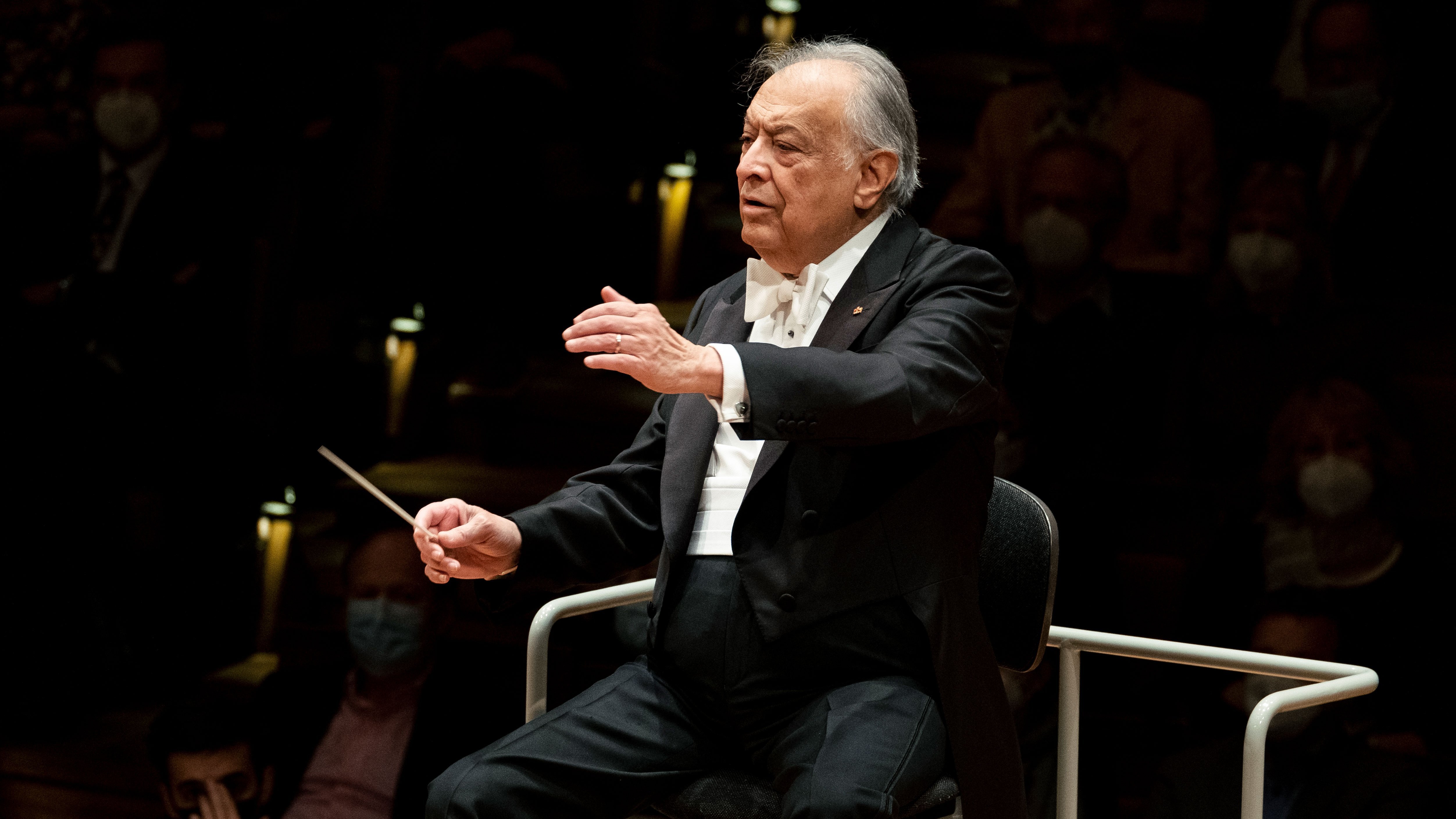 Ein älterer Dirigent in förmlicher Kleidung leitet ein Orchester im Sitzen und hält einen Taktstock in der Hand, während das Publikum im schwach beleuchteten Hintergrund verschwommen zu sehen ist.