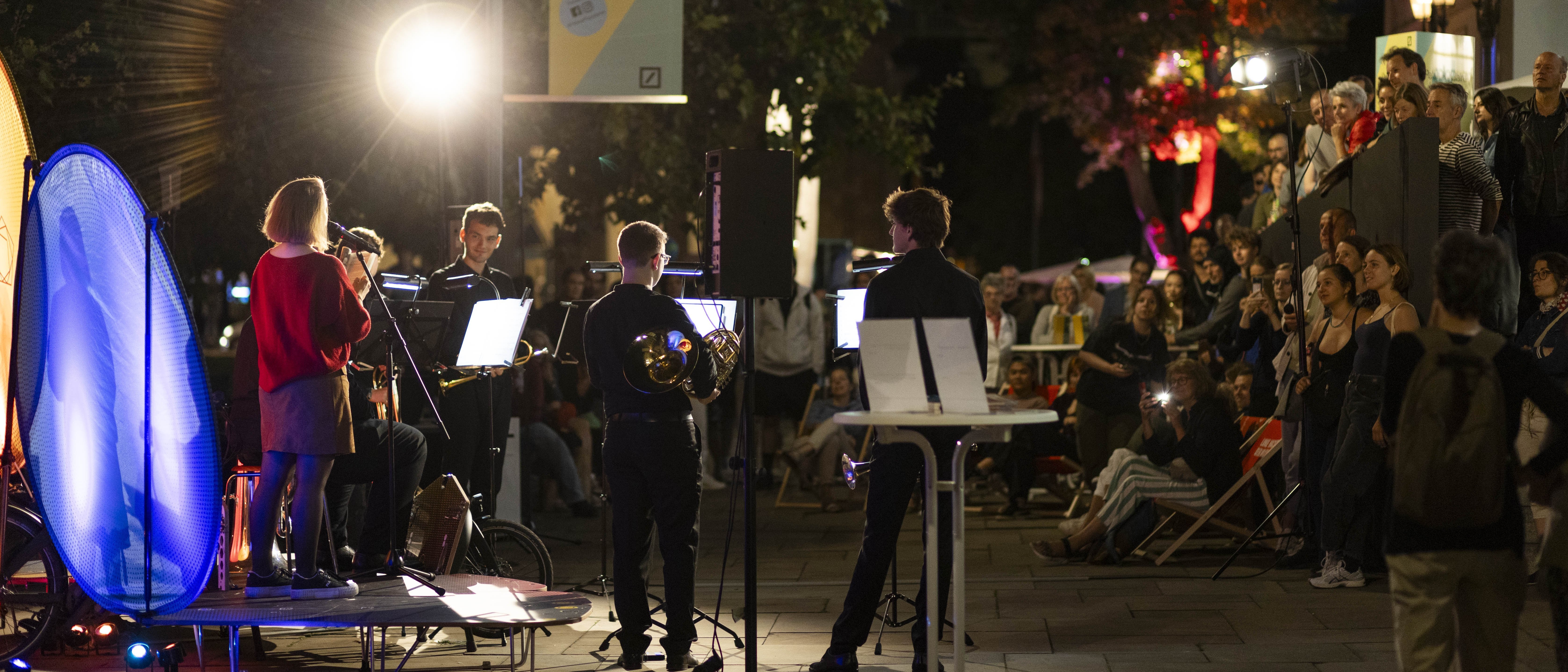 Ein kleines Musikensemble spielt nachts unter freiem Himmel vor einem sitzenden und stehenden Publikum auf einer städtischen Straße, beleuchtet von Bühnen- und Straßenlaternen. Auf einem Transparent darüber steht: "Kunst und Kultur entdecken".