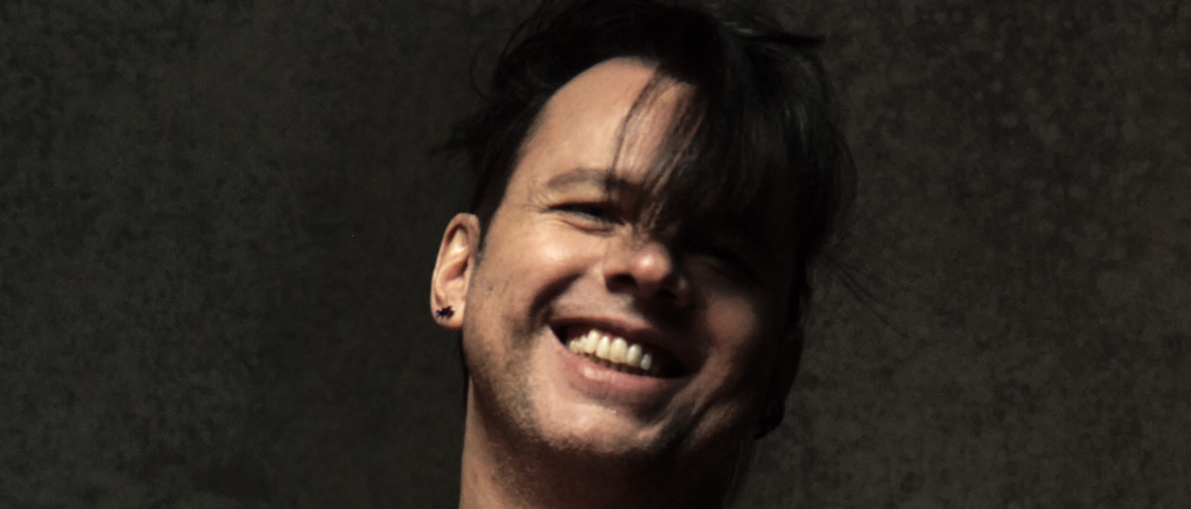 A person with short, dark hair and a side-swept fringe smiles broadly. They are wearing a sleeveless black top and small earring, standing against a textured dark background.