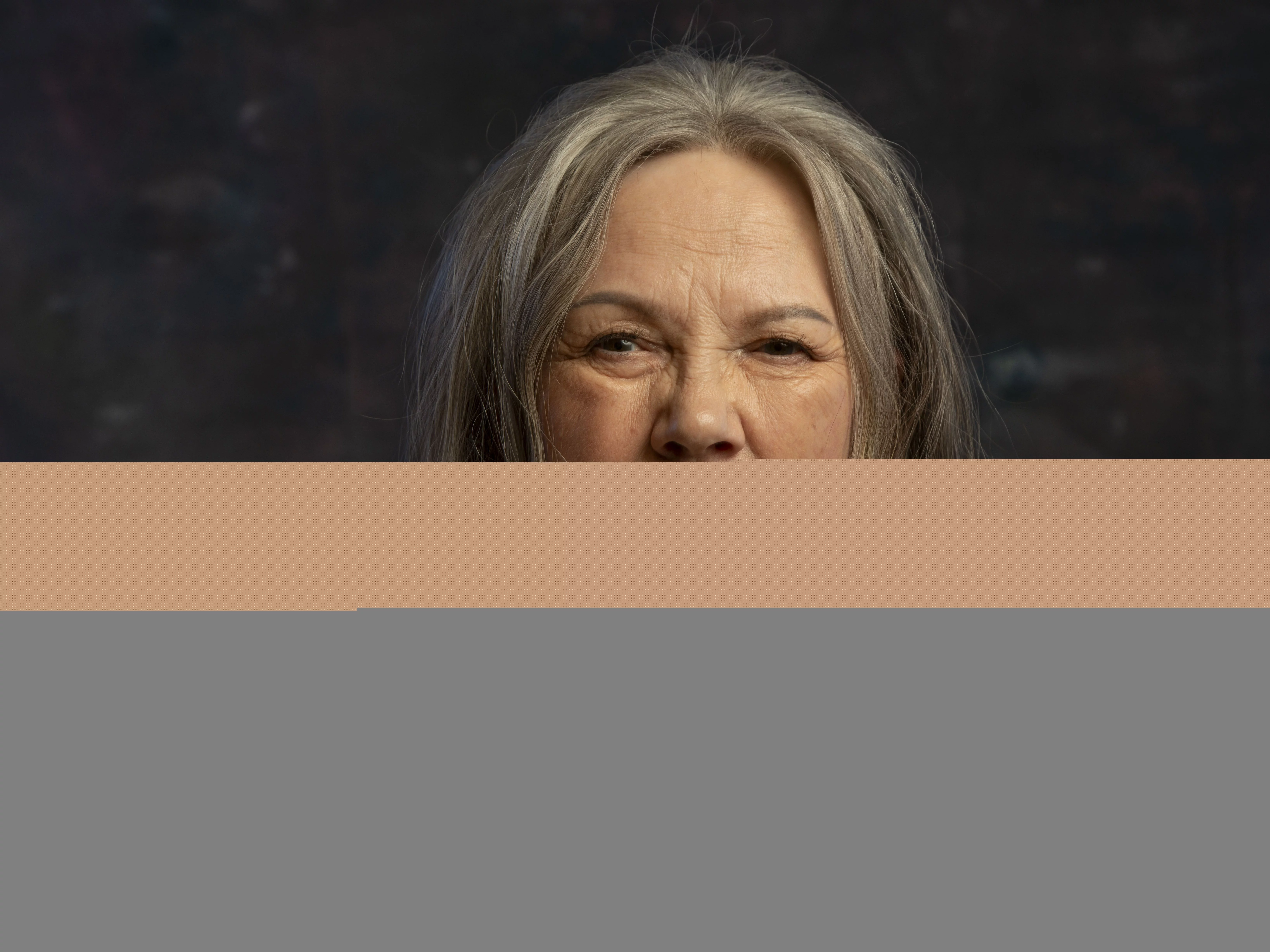 Mari Boine mit langem grauem Haar, roten Quastenohrringen und bunten Halsketten, in Schwarz gekleidet, vor einem dunklen, strukturierten Hintergrund.