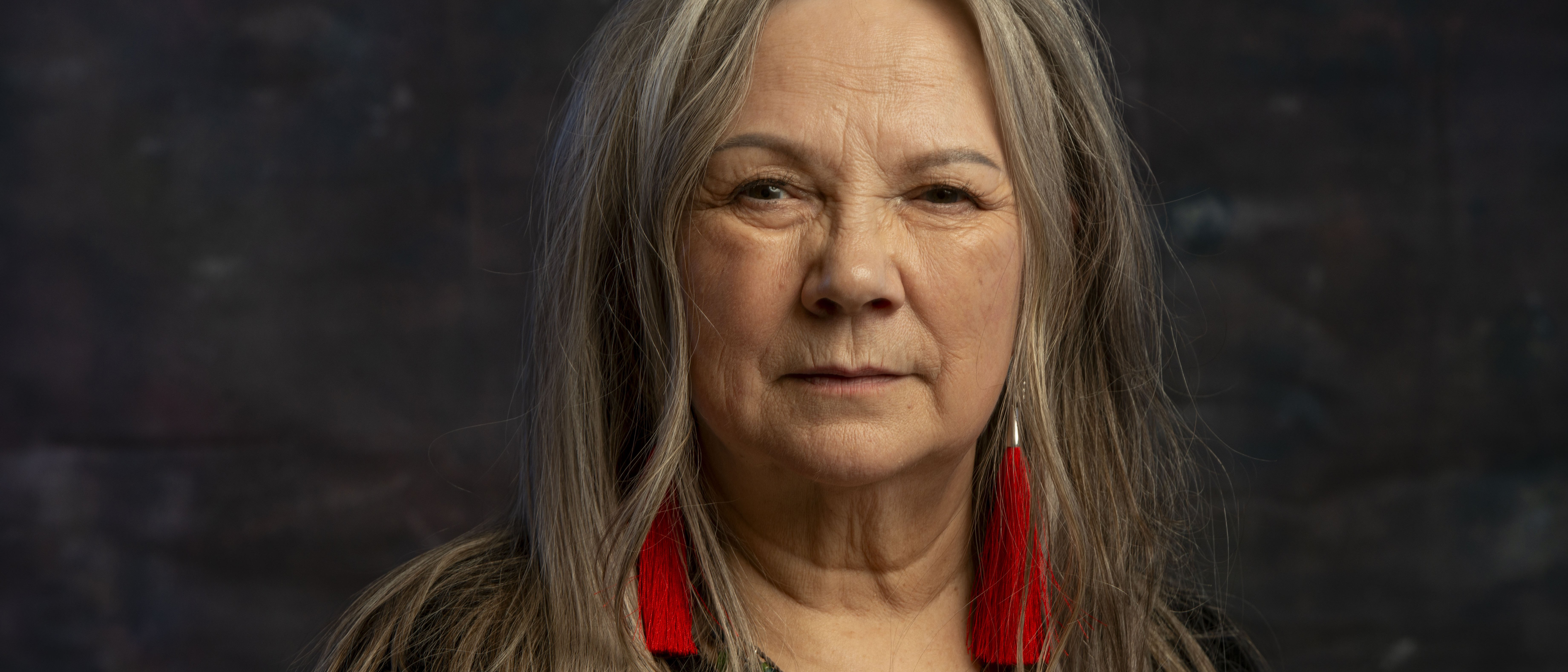 Mari Boine with long grey hair, red tassel earrings and colourful necklaces, dressed in black, against a dark, textured background.