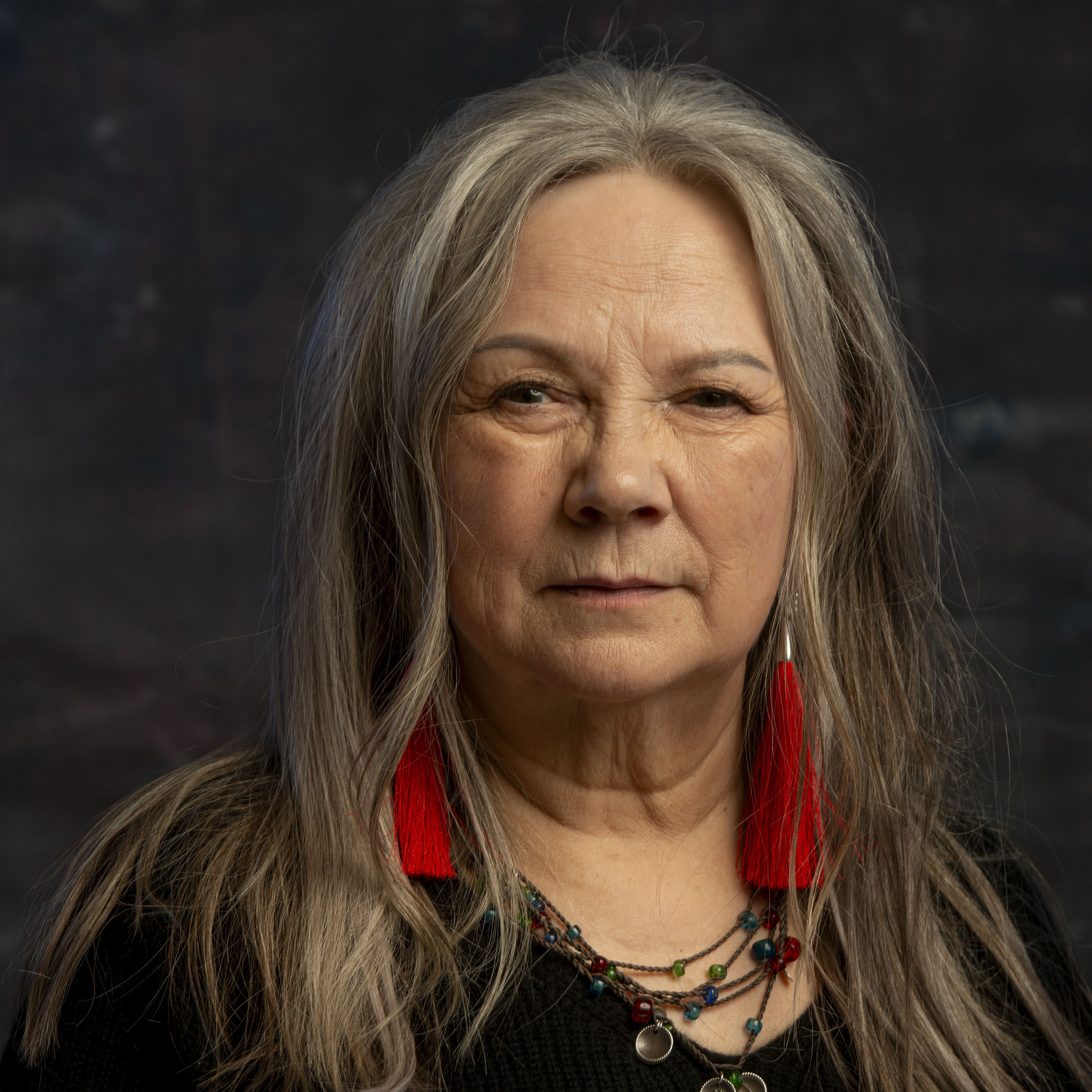 Mari Boine mit langem grauem Haar, roten Quastenohrringen und bunten Halsketten, in Schwarz gekleidet, vor einem dunklen, strukturierten Hintergrund.