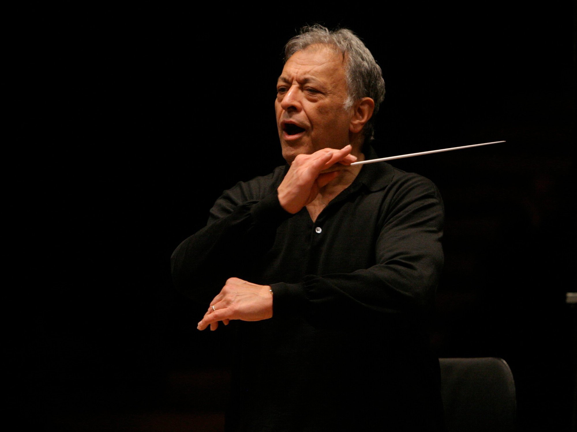 Ein älterer Mann in einem schwarzen Hemd dirigiert mit einem Taktstock, einem erhobenen Arm und offenem Mund, konzentriert wirkend, ein Orchester vor einem dunklen Hintergrund.