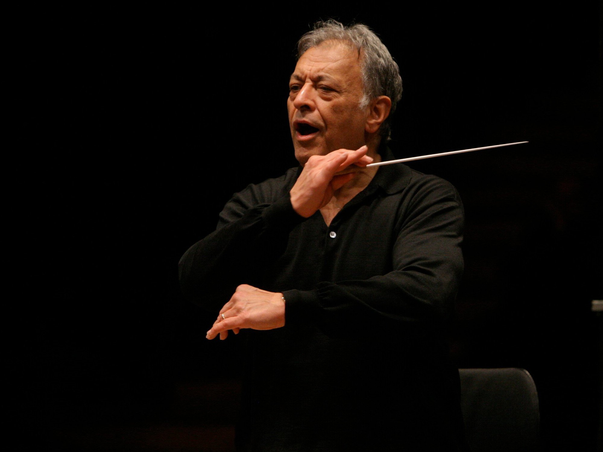 An older man wearing a black shirt conducts with a baton, his mouth open as if singing or giving instructions, set against a dark background.