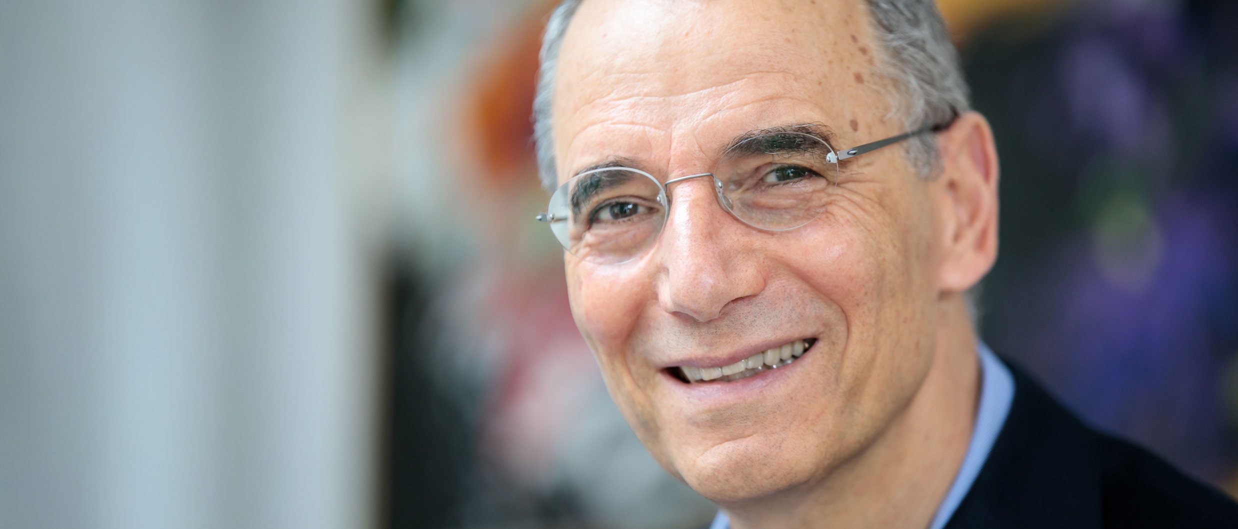 Ein lächelnder Mann mittleren Alters mit kurzen grauen Haaren und Brille, der eine dunkle Anzugsjacke und ein hellblaues Hemd trägt, posiert in einem Haus vor einem unscharfen, bunten Hintergrund.