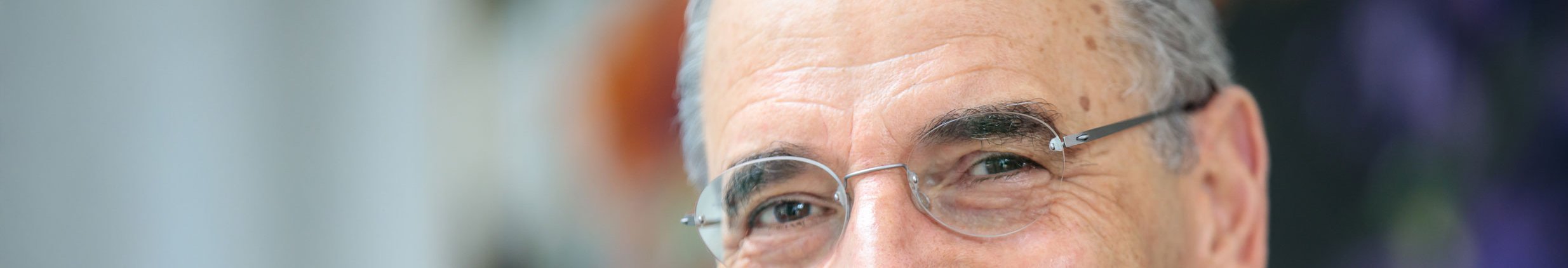 A middle-aged man wearing glasses and a suit jacket smiles warmly at the camera. The background is softly blurred with hints of colorful shapes.