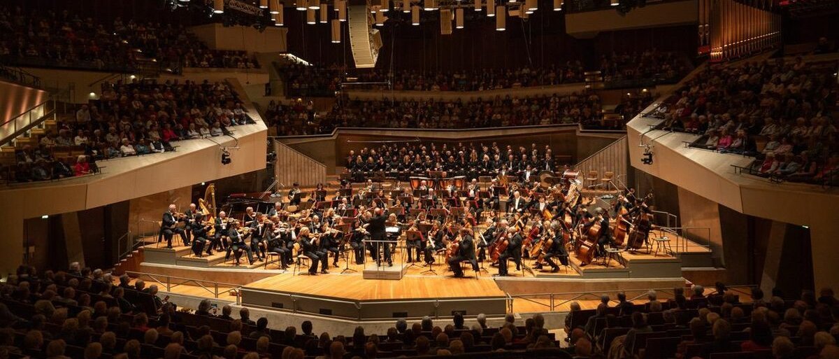 Ein vollbesetztes Orchester spielt auf der Bühne in einem großen, modernen Konzertsaal, der mit Zuschauern gefüllt ist. Hinter dem Orchester stehen Chormitglieder, und eine warme Beleuchtung taucht die Szene in ein warmes Licht.