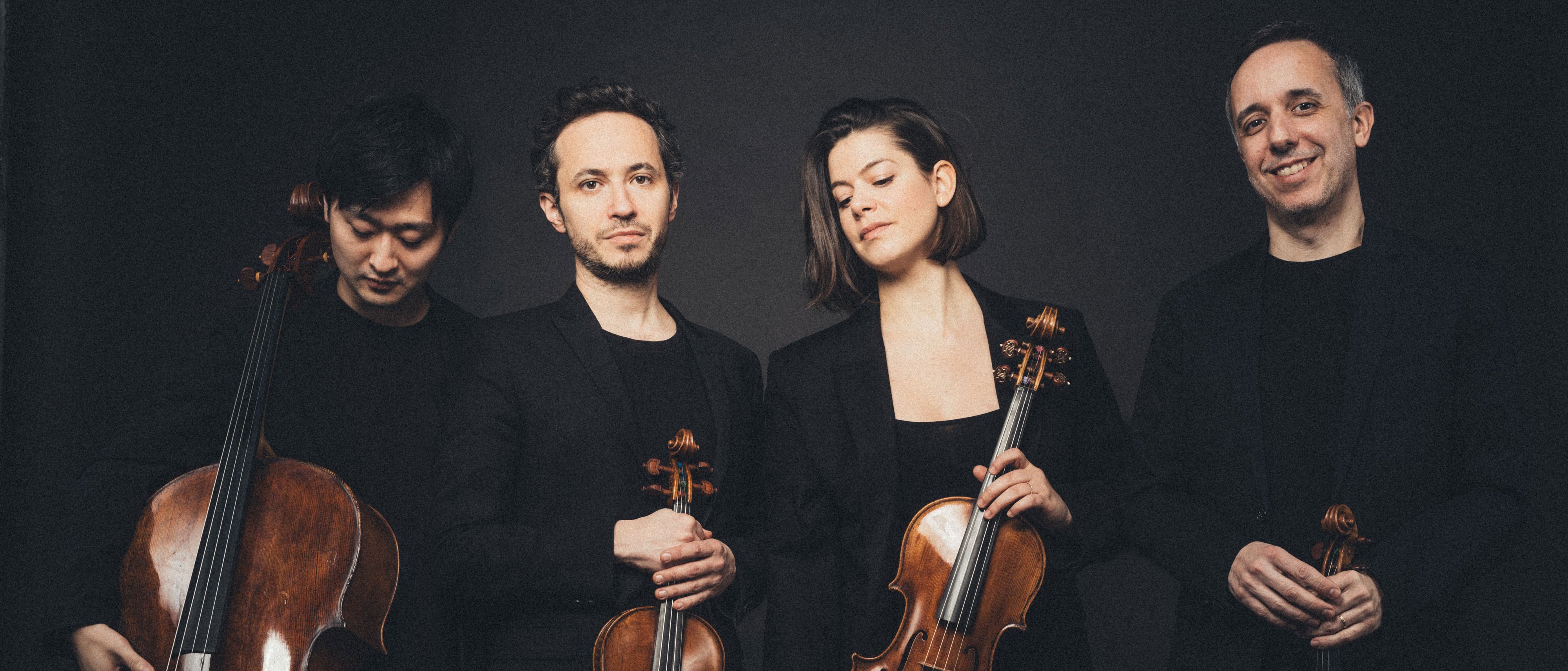 Vier Musiker posieren mit Streichinstrumenten vor dunklem Hintergrund. Sie tragen schwarze Abendgarderobe und halten ein Cello, zwei Geigen und eine Bratsche in den Händen. Sie stehen eng beieinander und blicken mit entspanntem Gesichtsausdruck in die Kamera.