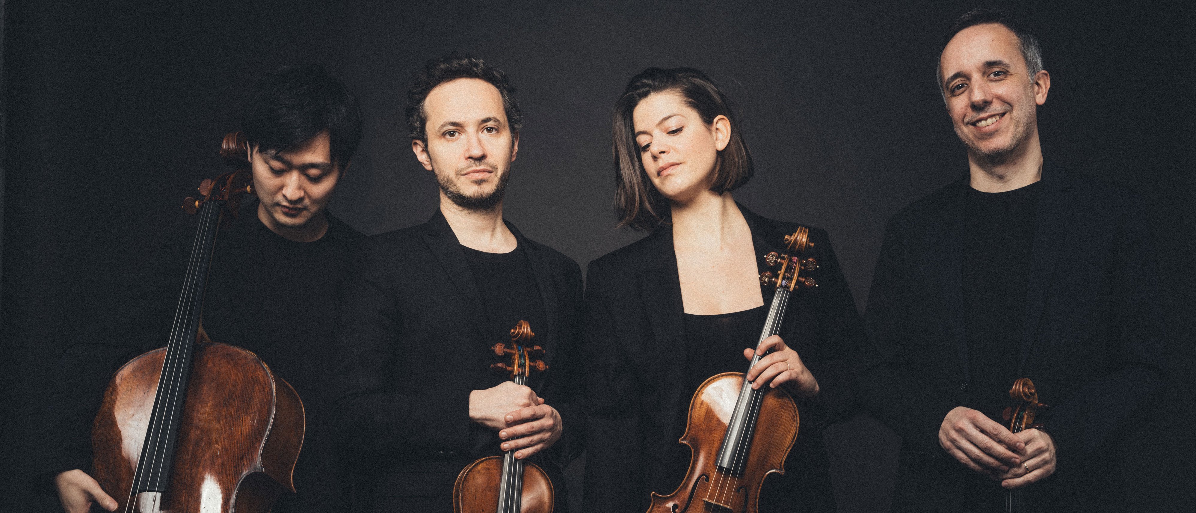Four musicians pose with string instruments in front of a dark background. They wear black evening dress and hold a cello, two violins and a viola in their hands. They stand close together and look at the camera with a relaxed expression on their faces.