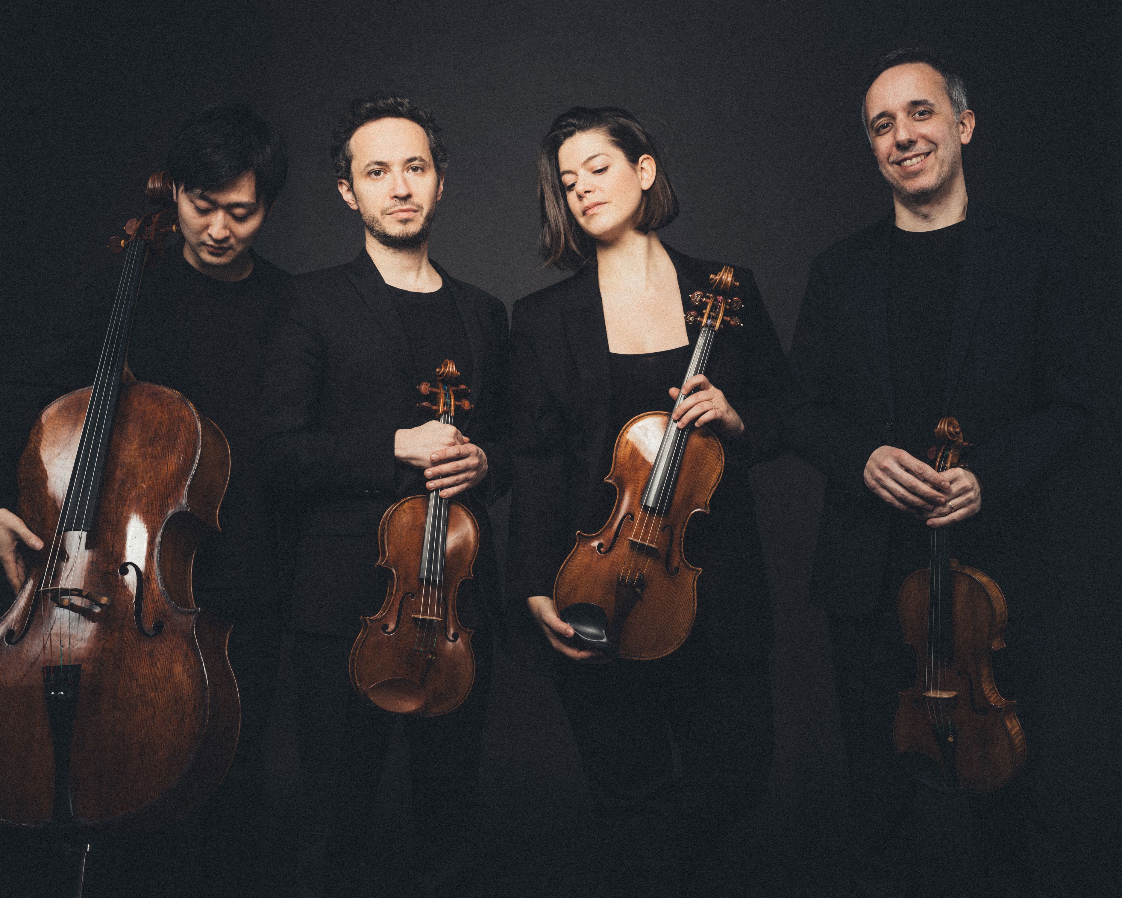 Vier Musiker posieren mit Streichinstrumenten vor dunklem Hintergrund. Sie tragen schwarze Abendgarderobe und halten ein Cello, zwei Geigen und eine Bratsche in den Händen. Sie stehen eng beieinander und blicken mit entspanntem Gesichtsausdruck in die Kamera.