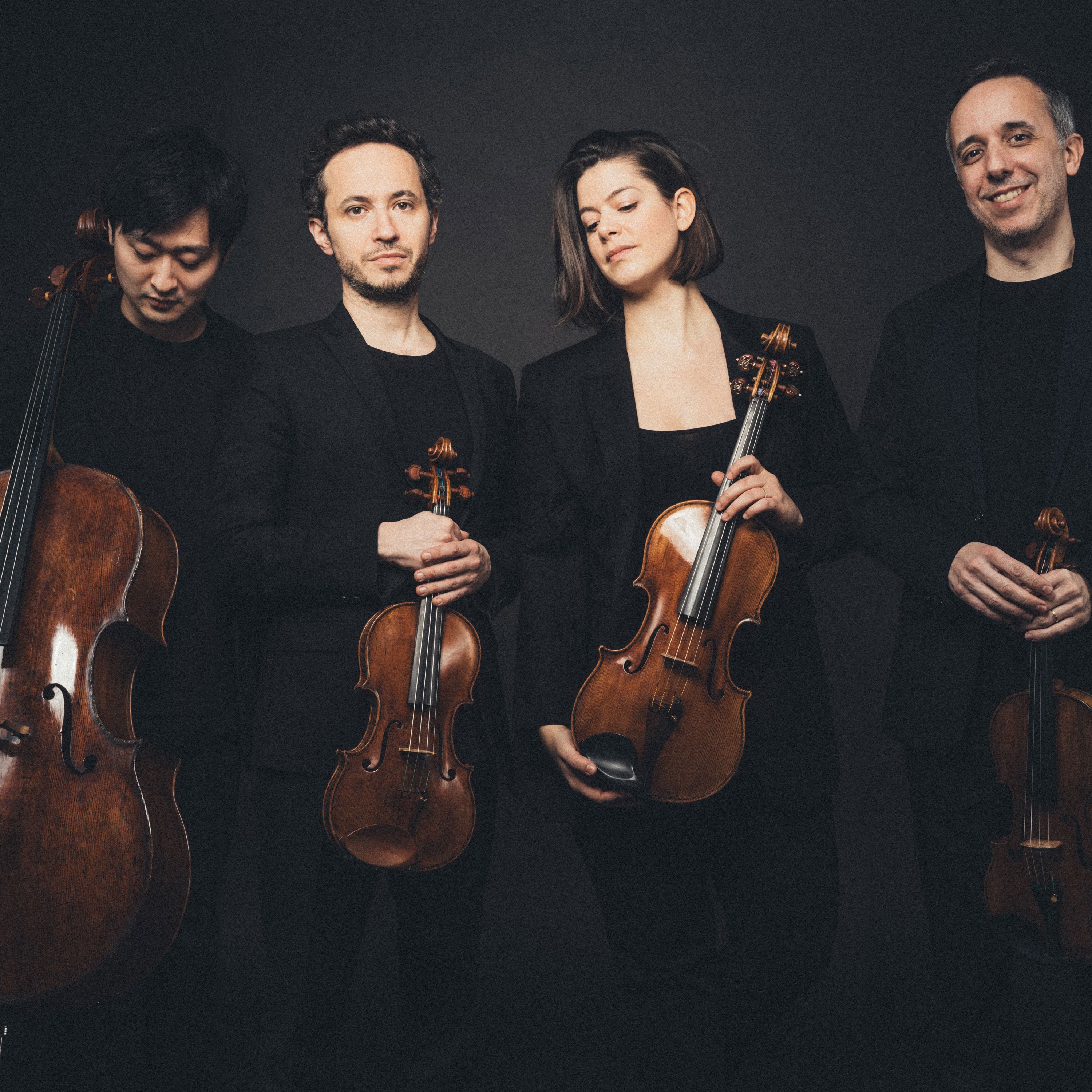 Vier Musiker posieren mit Streichinstrumenten vor dunklem Hintergrund. Sie tragen schwarze Abendgarderobe und halten ein Cello, zwei Geigen und eine Bratsche in den Händen. Sie stehen eng beieinander und blicken mit entspanntem Gesichtsausdruck in die Kamera.