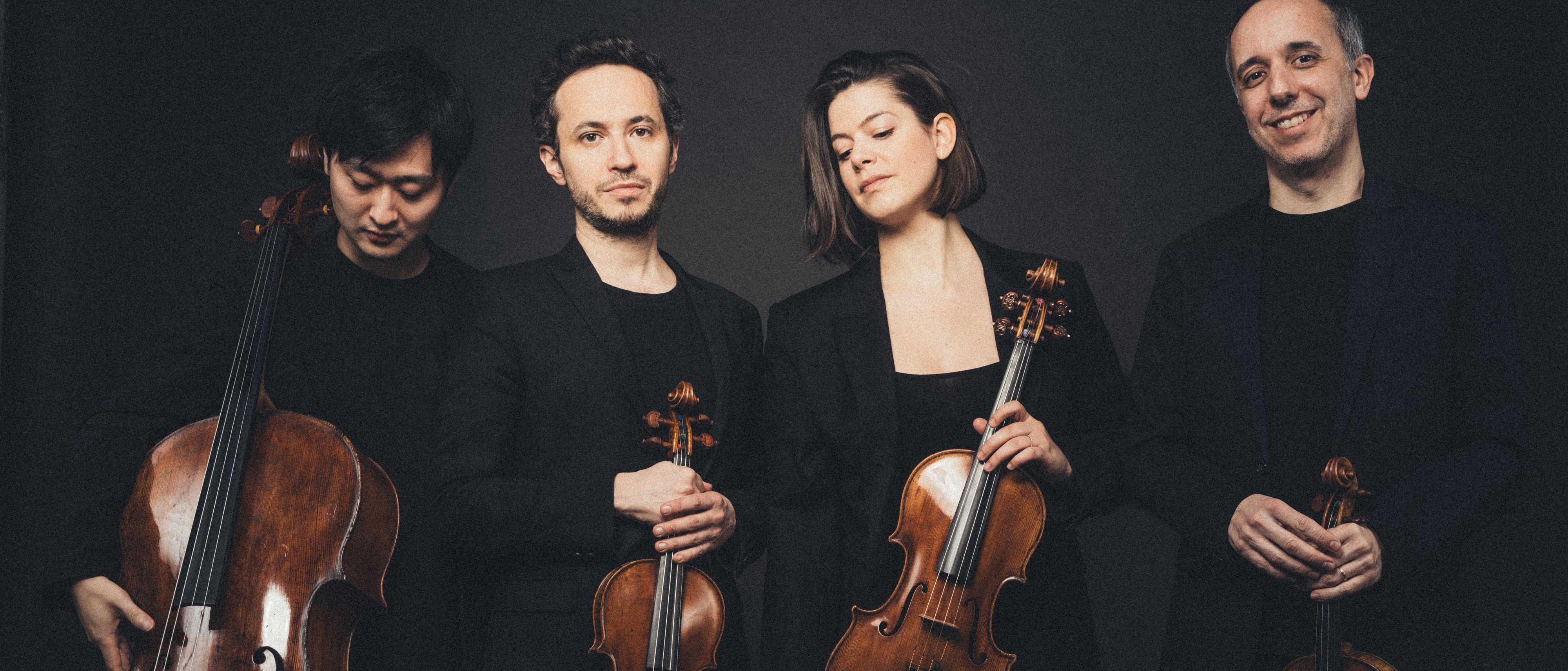 Vier Musiker posieren mit Streichinstrumenten vor dunklem Hintergrund. Sie tragen schwarze Abendgarderobe und halten ein Cello, zwei Geigen und eine Bratsche in den Händen. Sie stehen eng beieinander und blicken mit entspanntem Gesichtsausdruck in die Kamera.