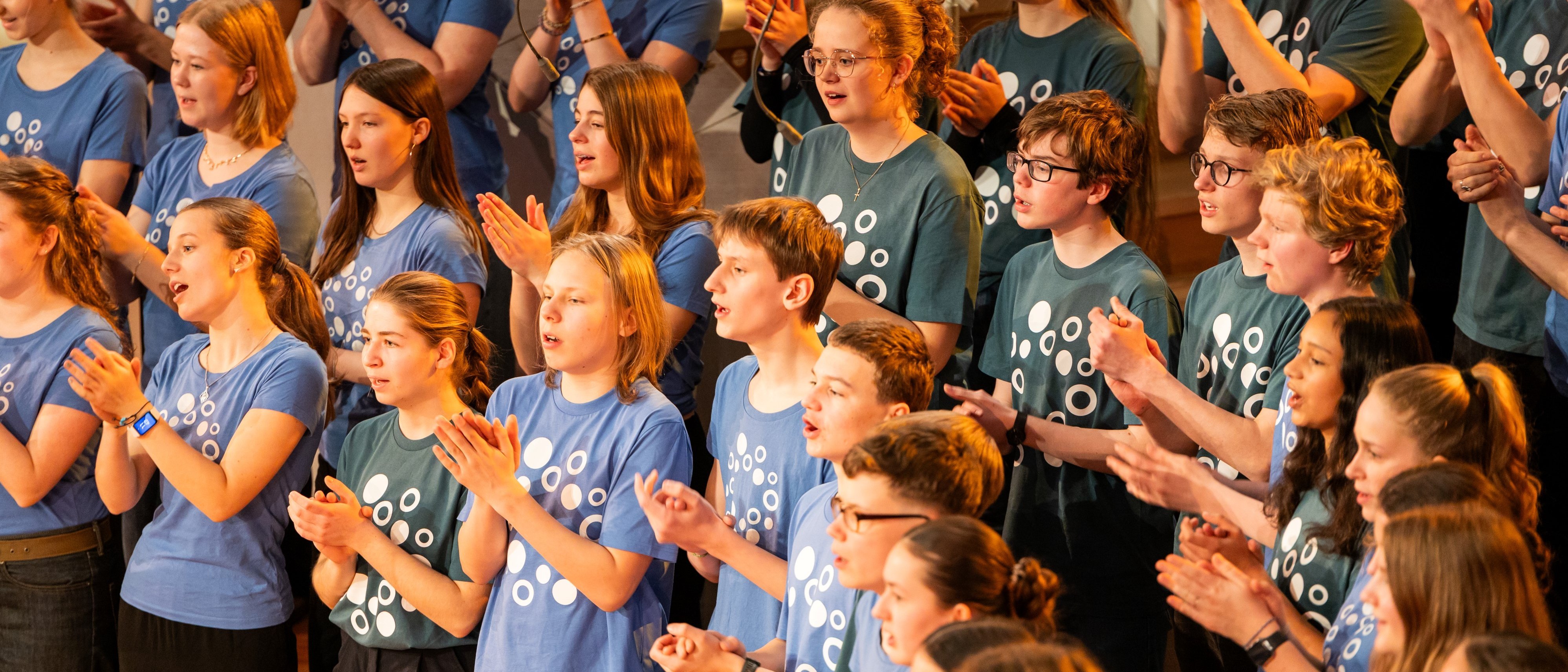 Eine Gruppe junger Leute in passenden blauen Hemden singt und klatscht begeistert im Chor. Im Hintergrund sind weitere Mitglieder zu sehen, die sich der Darbietung anschließen. Sie stehen auf der Bühne und drücken Freude und Energie aus.