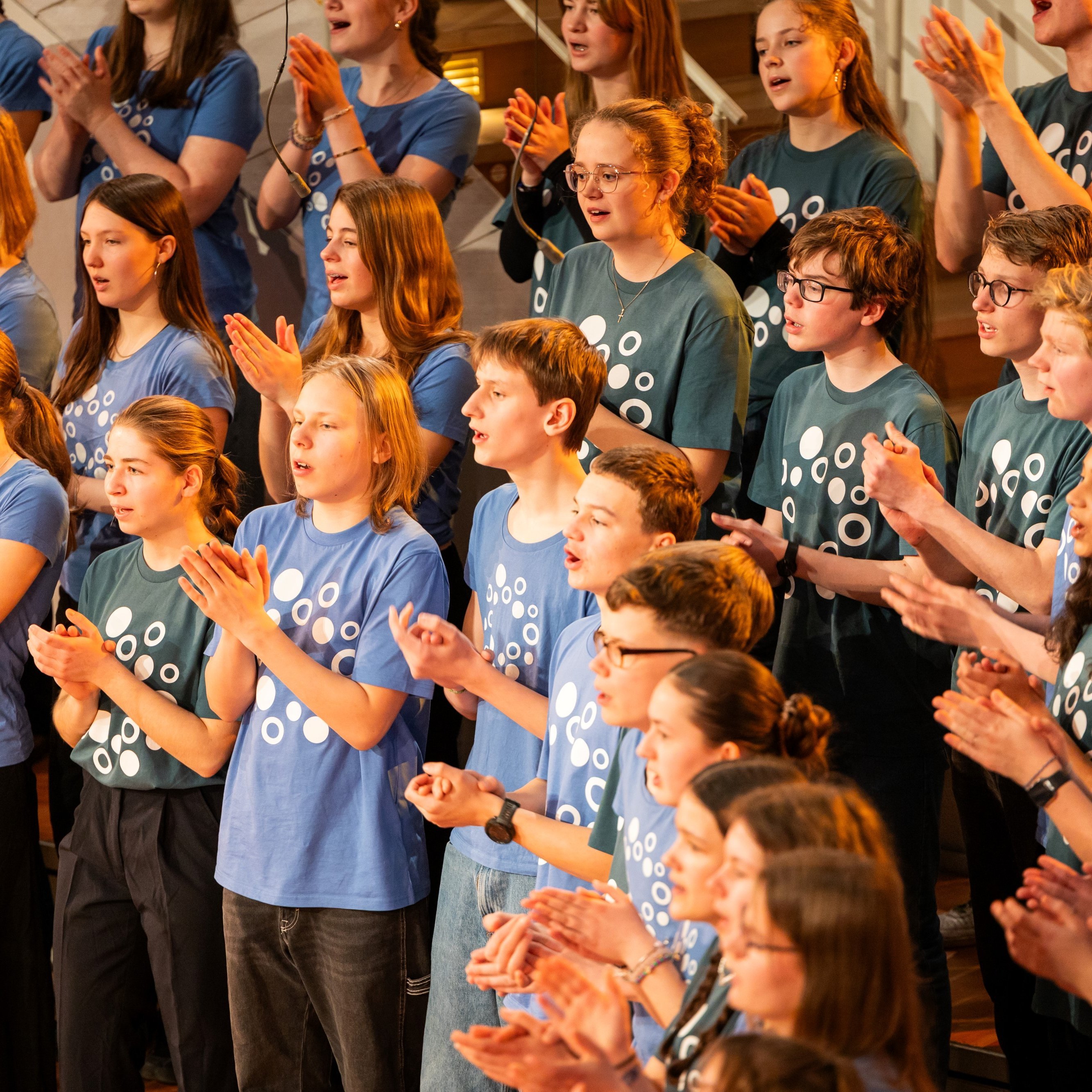 Eine Gruppe junger Leute in passenden blauen Hemden singt und klatscht begeistert im Chor. Im Hintergrund sind weitere Mitglieder zu sehen, die sich der Darbietung anschließen. Sie stehen auf der Bühne und drücken Freude und Energie aus.