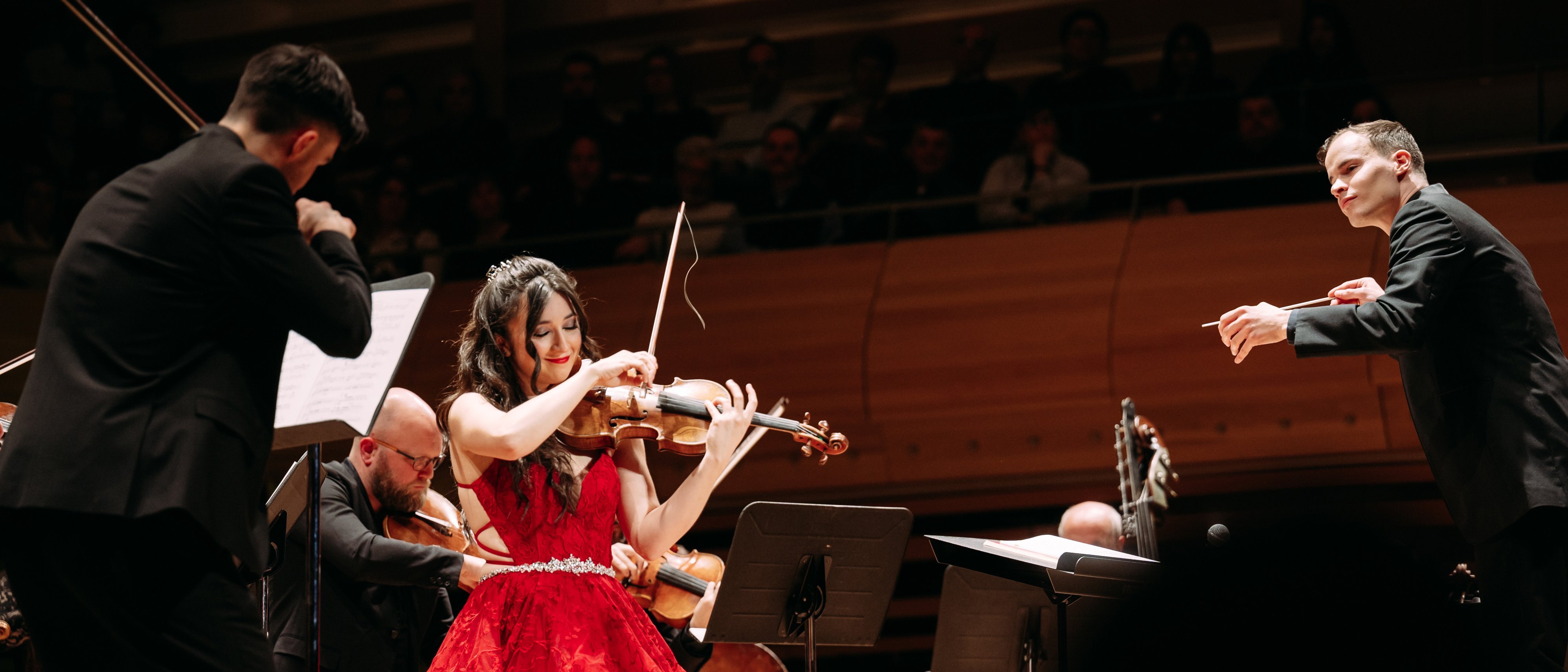 Eine Frau in einem leuchtend roten Abendkleid spielt auf der Bühne zusammen mit einem Orchester Geige. Zu sehen sind außerdem Musiker in schwarzer Abendgarderobe und ein Dirigent, während im Hintergrund das Publikum zuschaut.
