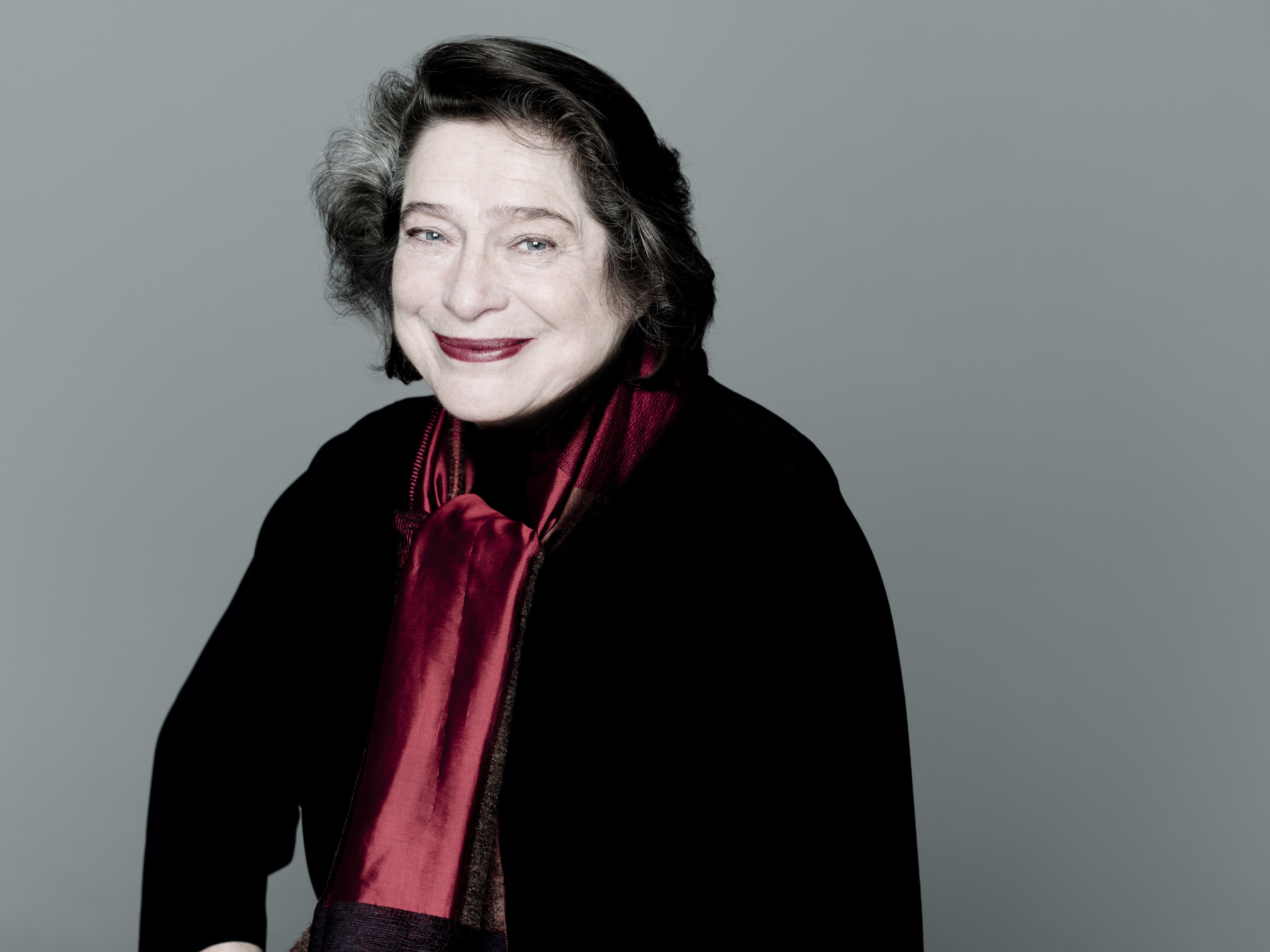 Sitzende Elisabeth Leonskaja, leicht lächelnd, mit dunklem Haar. Sie trägt ein dunkles Outfit und einen roten Schal vor einem schlichten grauen Hintergrund.
