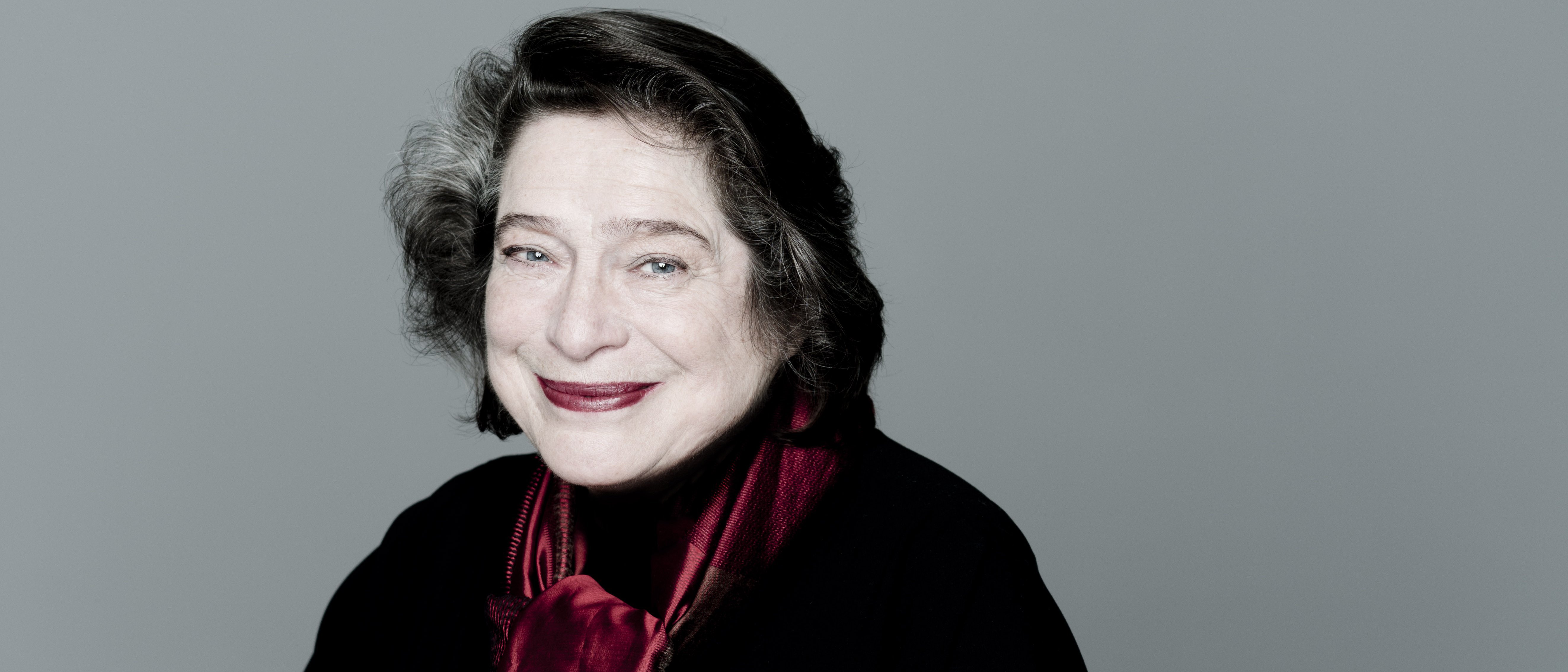 Elisabeth Leonskaja, seated and gently smiling, with dark hair. She wears a dark outfit and a red scarf, set against a plain grey background.