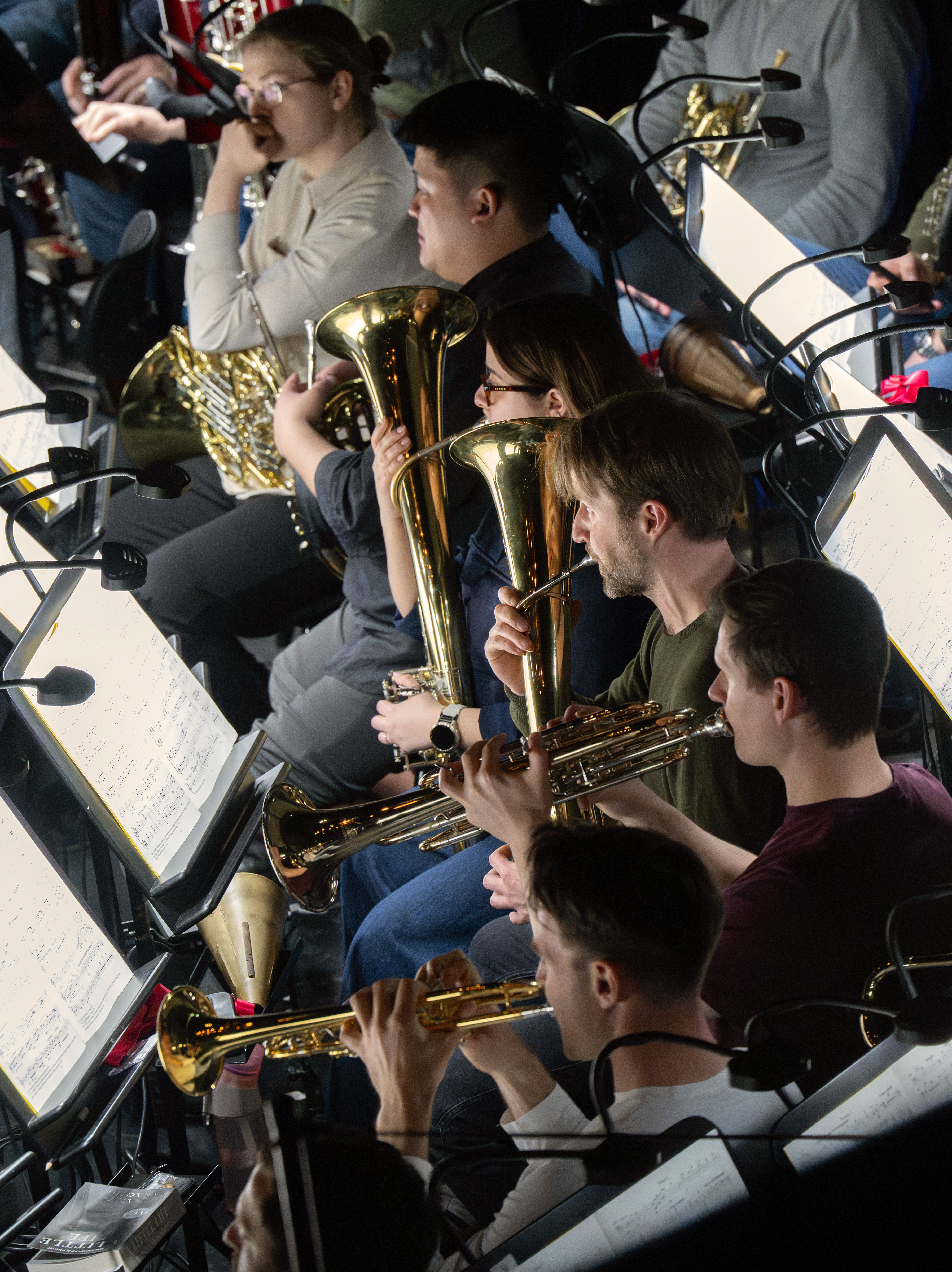Mehrere Musiker sitzen eng beieinander in einem Orchestergraben, halten Blechblasinstrumente in der Hand und lesen Notenblätter auf Ständern, um sich auf ihr Spiel vorzubereiten.