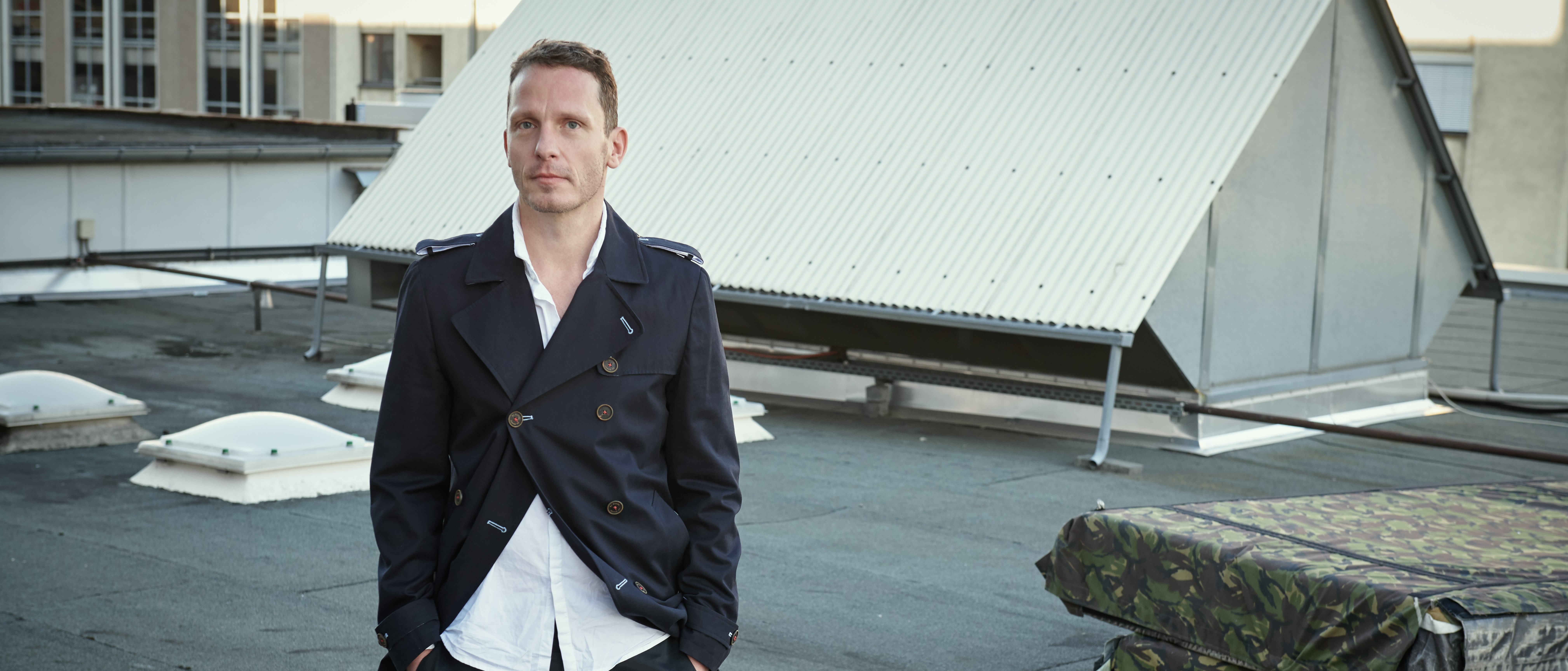 A man in a dark coat and white shirt stands on a rooftop with industrial structures and a stack of camouflage-covered objects in the background. He has his hands in his pockets and looks ahead.