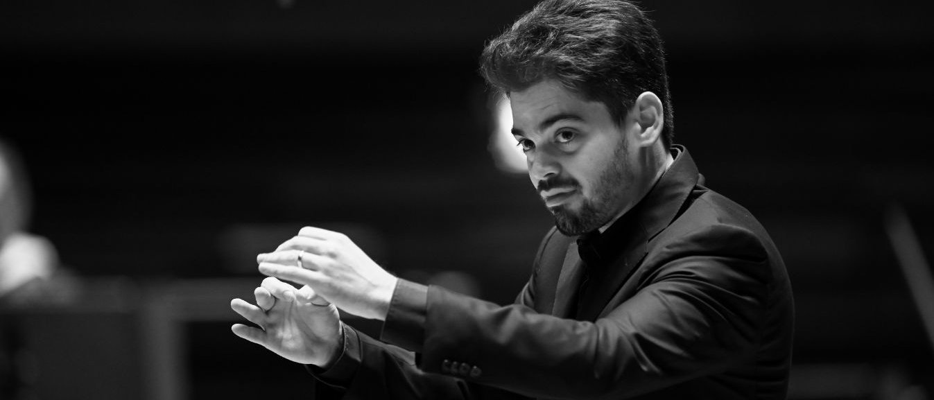 Lahav Shani im Anzug dirigiert ein Orchester mit erhobenen Händen und konzentriertem Gesichtsausdruck, mit unscharfen Musikern und Instrumenten im Hintergrund. Schwarz-Weiß-Foto.