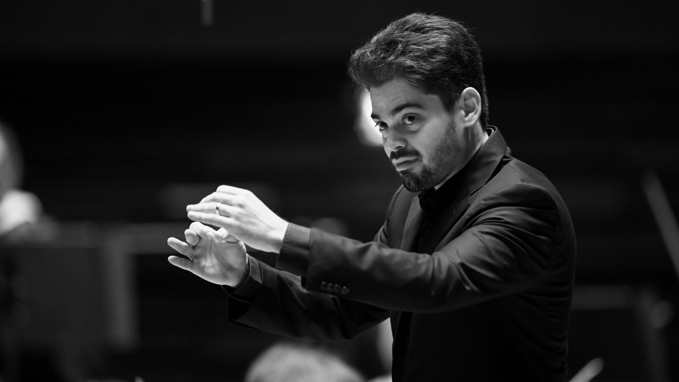 Lahav Shani in a dark suit conducts an orchestra, holding his hands mid-air with a focused expression. The background is blurred, emphasizing his gesture and facial concentration. The image is in black and white.