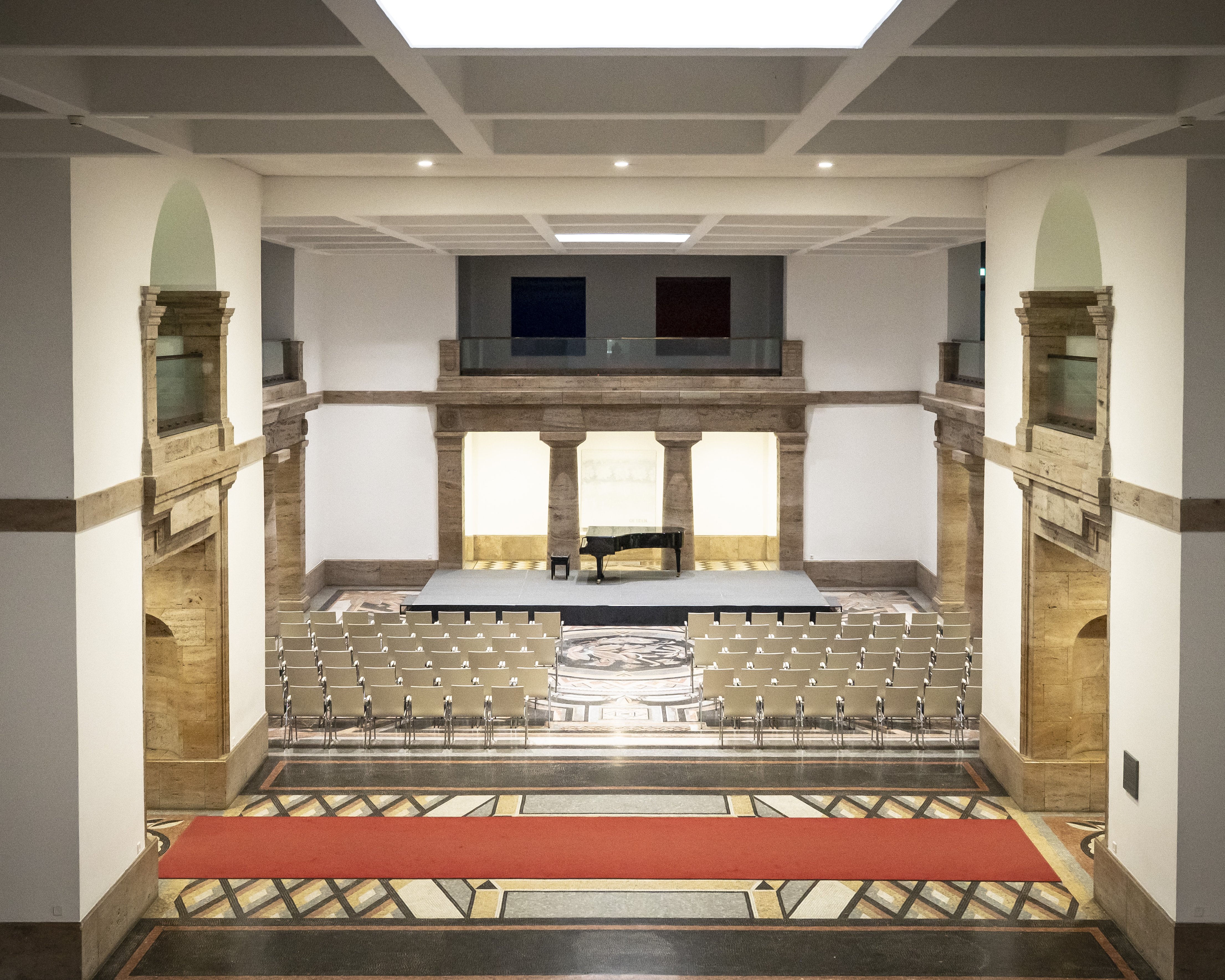 Blick in eine großzügige Mosaikhalle mit historischer Architektur, einem Flügel auf einer kleinen Bühne und davor aufgestellten Stuhlreihen.