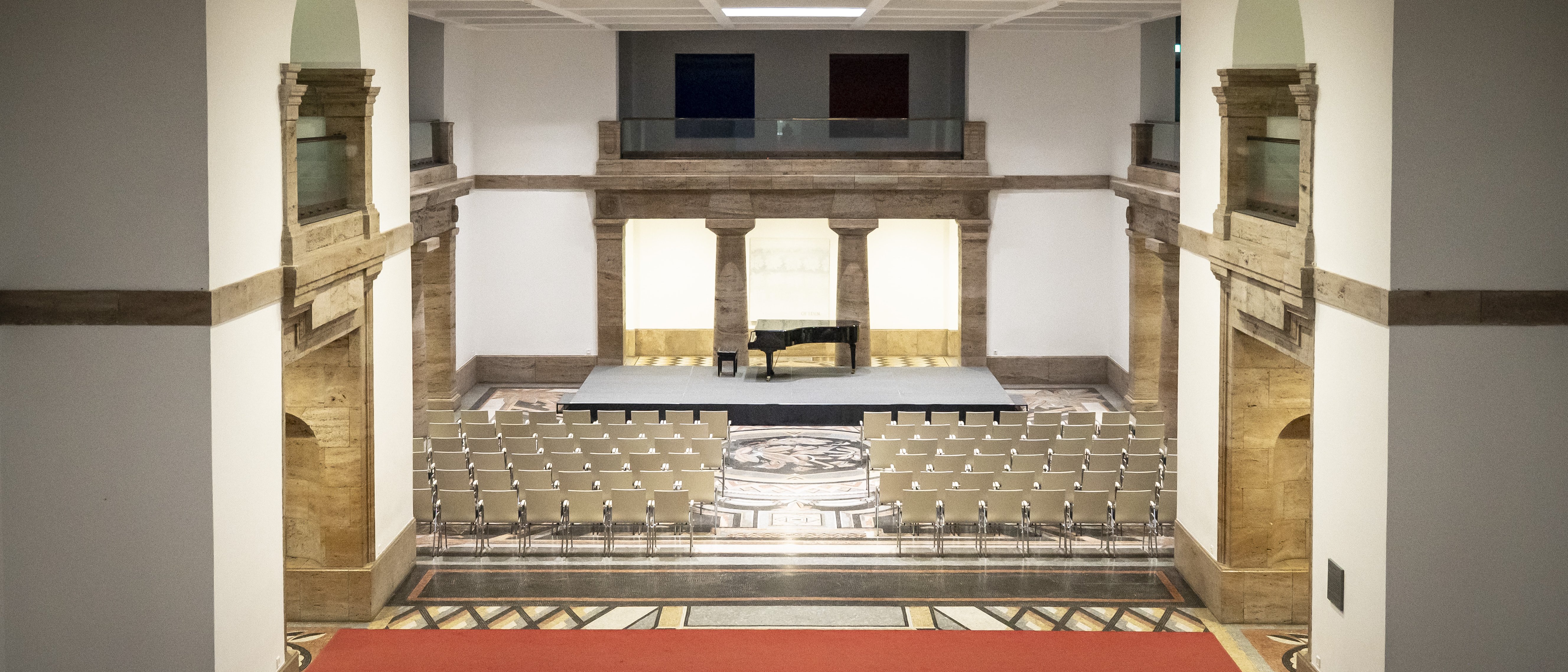 View into a spacious mosaic hall with historic architectural features, a grand piano on a small stage and rows of chairs arranged in front of it.
