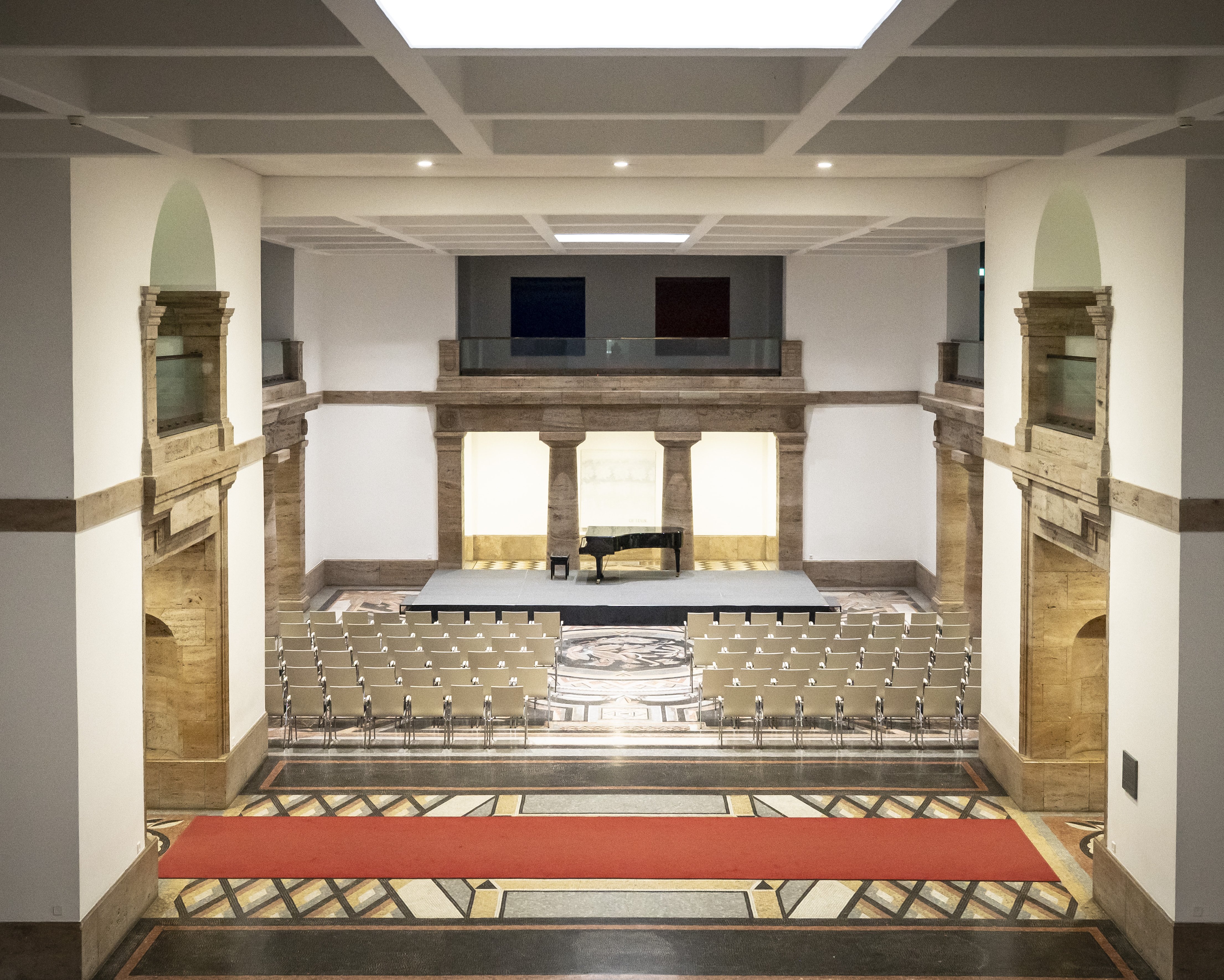 View into a spacious mosaic hall with historic architectural features, a grand piano on a small stage and rows of chairs arranged in front of it.