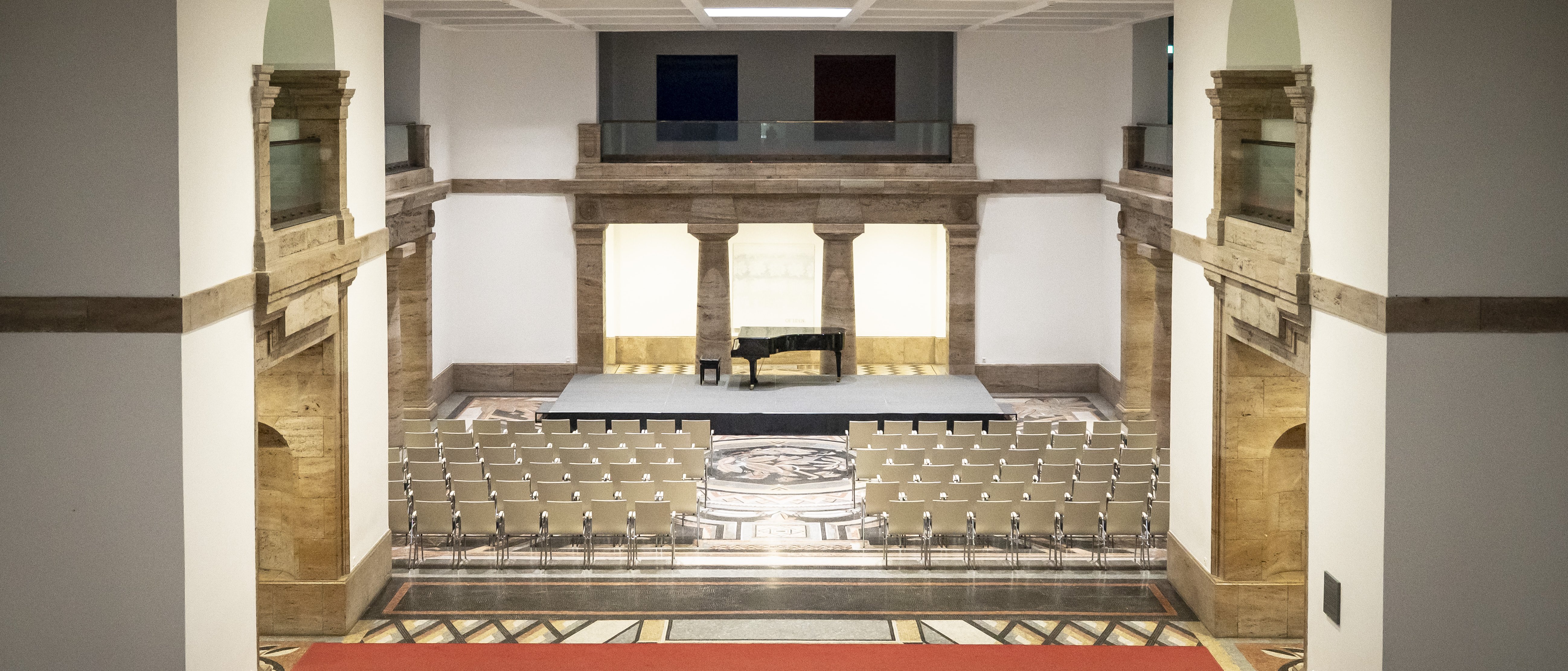 Blick in eine großzügige Mosaikhalle mit historischer Architektur, einem Flügel auf einer kleinen Bühne und davor aufgestellten Stuhlreihen.
