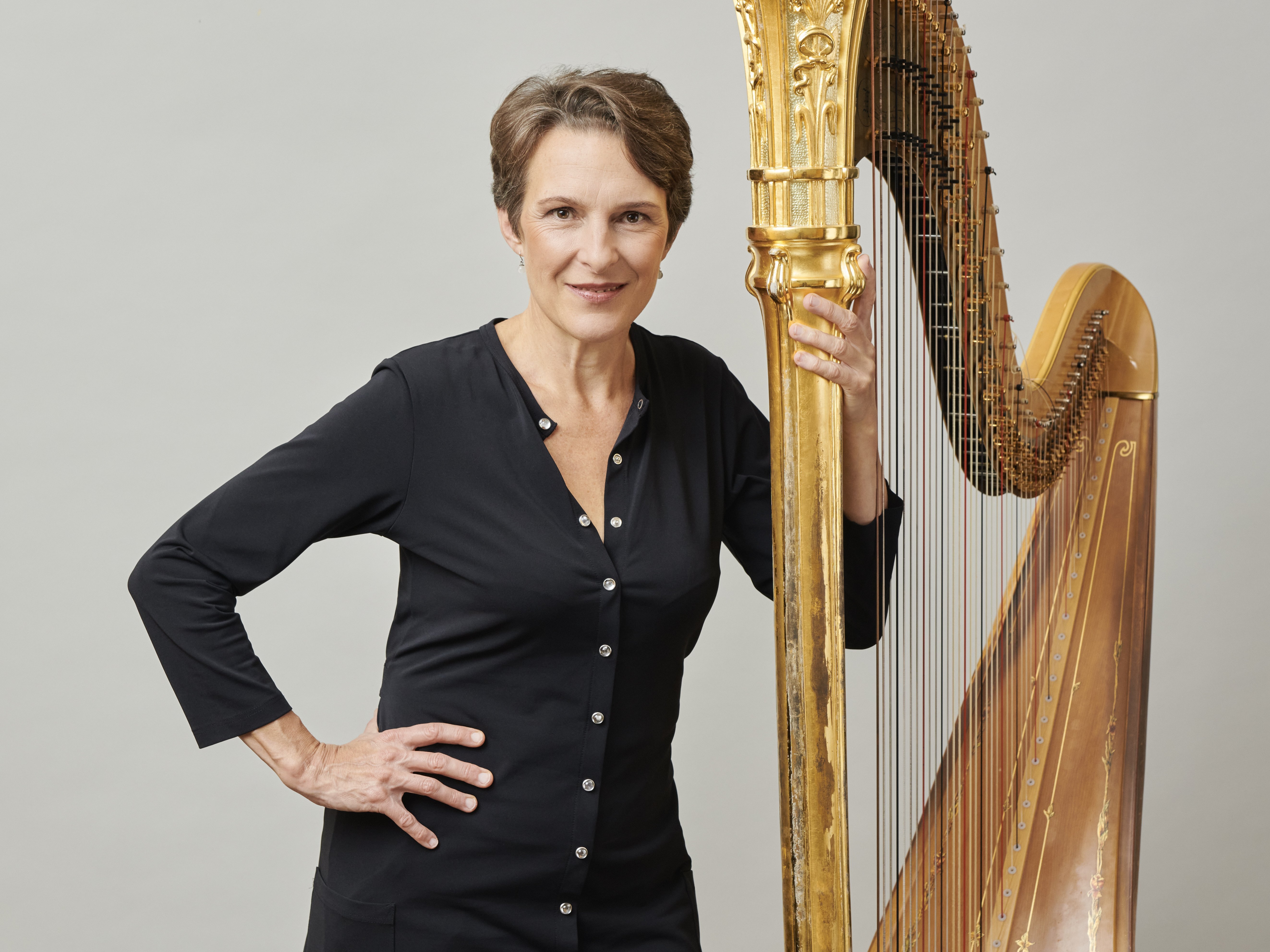 Marie-Pierre Langlamet steht vor einem grauen Hintergrund, mit der linken Hand hält sie ihre Harfe, die rechte hat sie in die Hüfte gestemmt.