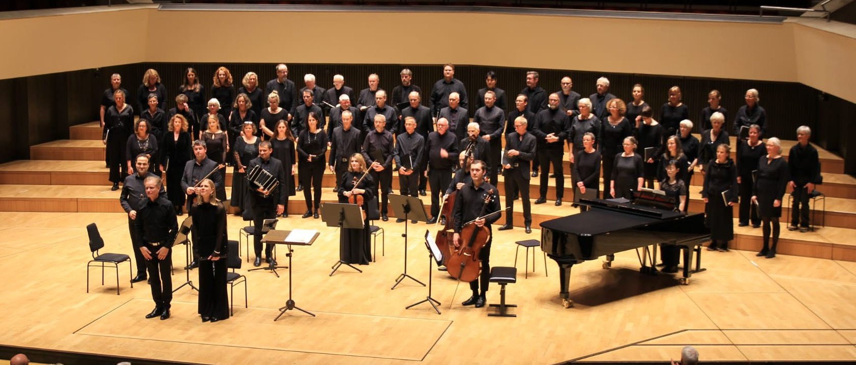 Ein Chor und Musiker stehen auf der Bühne eines Konzertsaals, ganz in Schwarz gekleidet; in ihrer Nähe stehen ein Flügel, Notenständer und Stühle, und sie blicken auf ein nicht zu seendes Publikum.