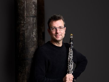 Ein Mann mit Brille und schwarzem Pullover steht vor einer dunklen Säule, hält eine Klarinette in der Hand und lächelt leicht, während der Hintergrund dunkelgrau ist.